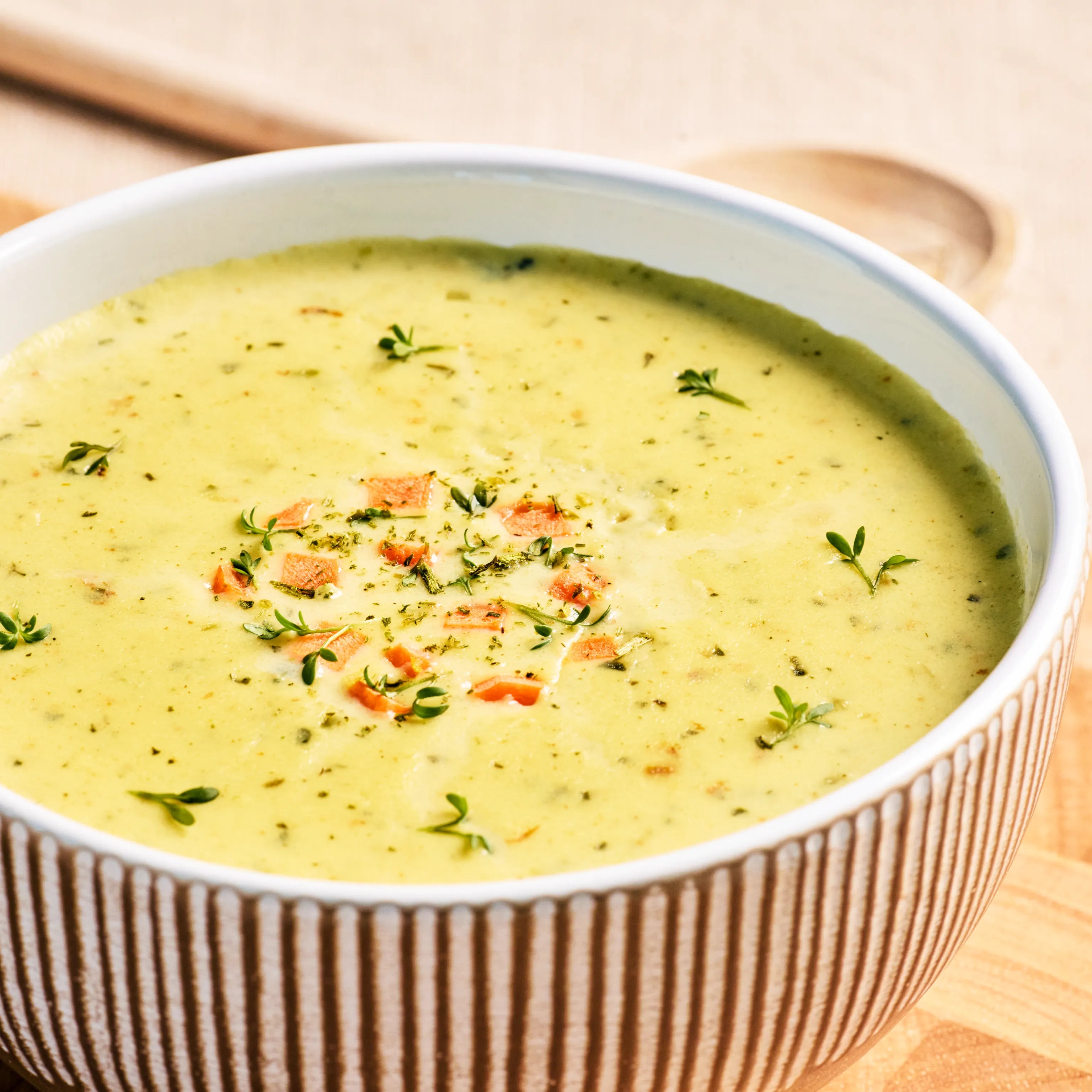 Schüssel mit cremiger Gemüsesuppe, garniert mit Kräutern und kleinen Karottenstücken, auf hellem Holztisch