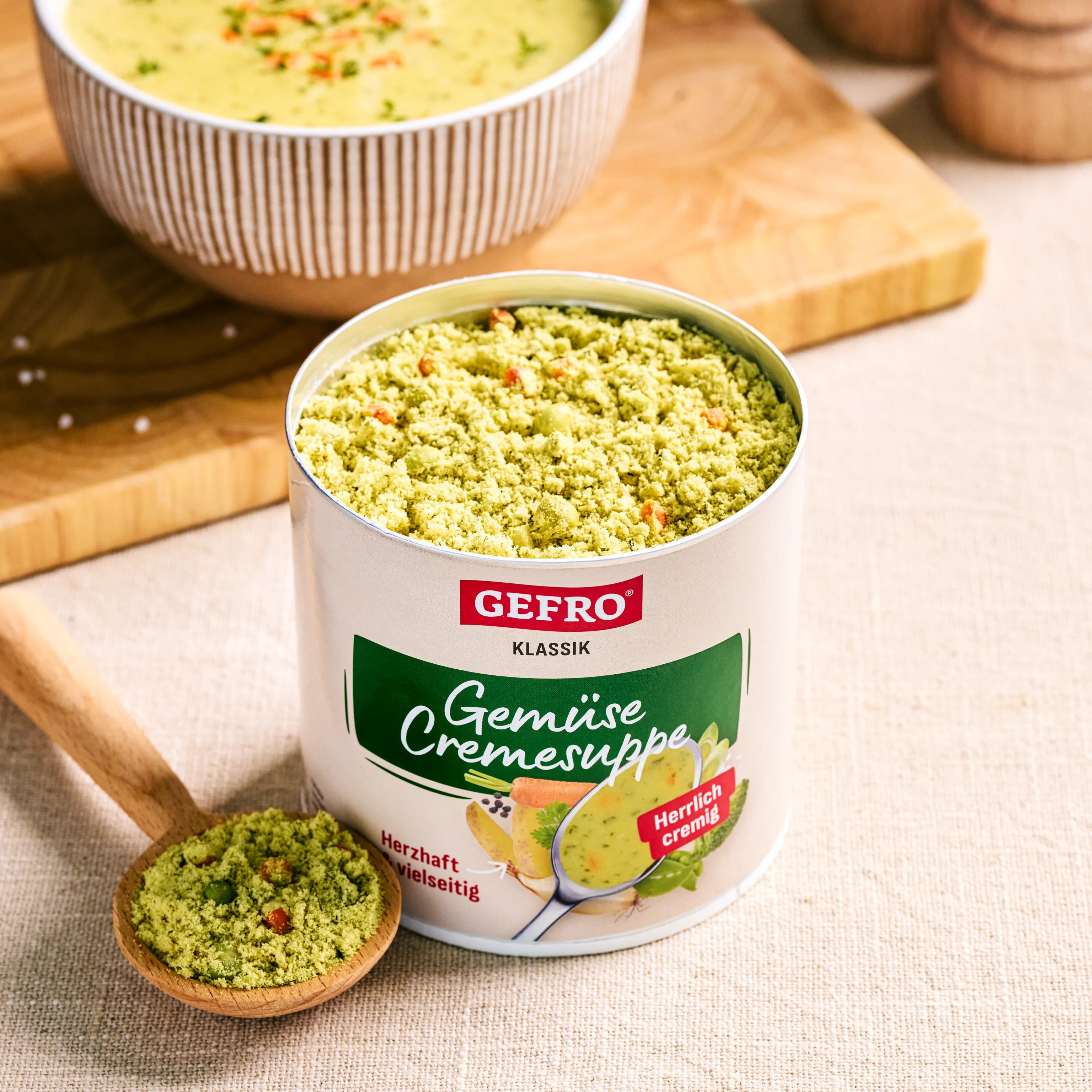 Dose GEFRO Klassik Gemüse Cremesuppe mit geöffnetem Pulverinhalt auf Tisch, daneben Löffel und zubereitete Suppe