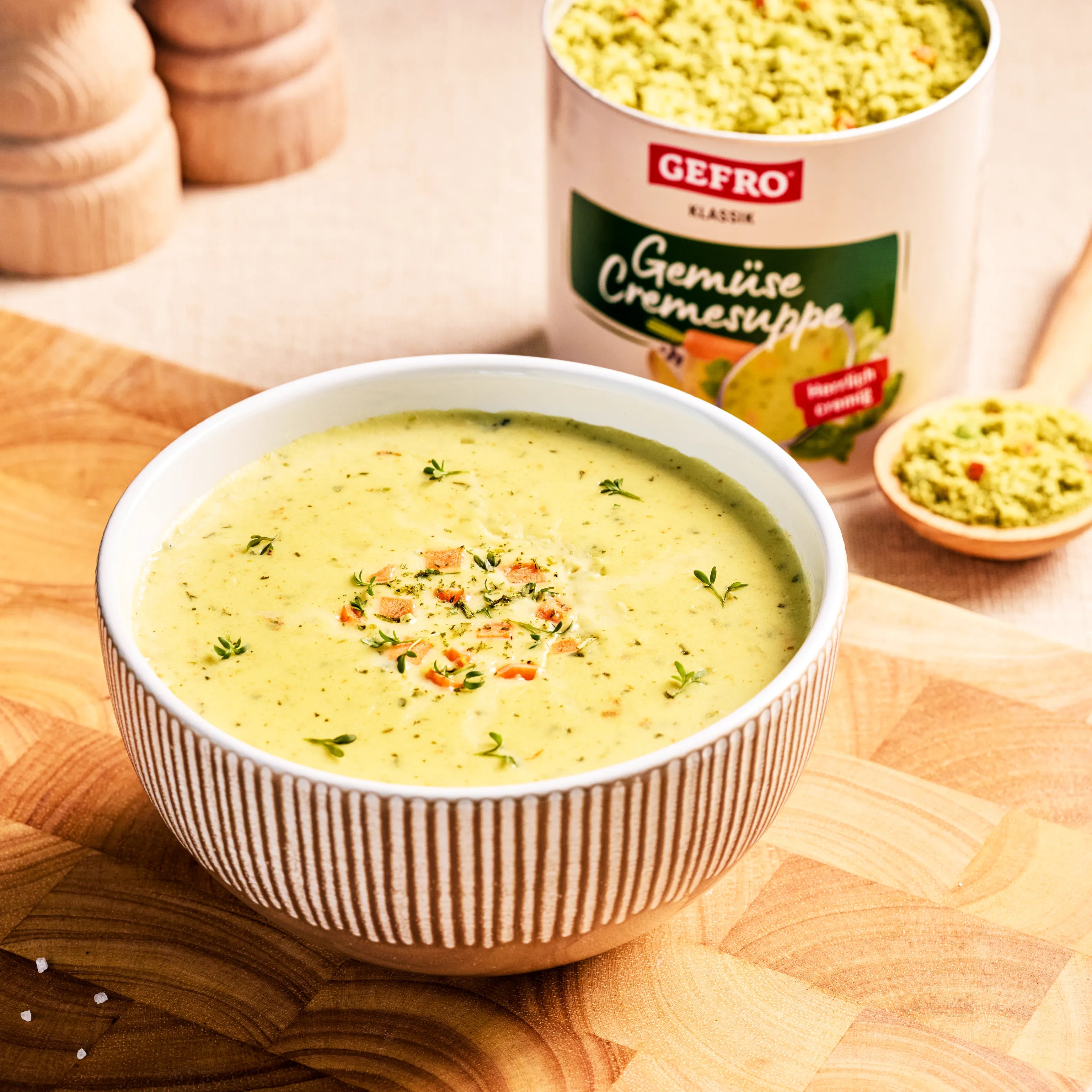 Schale mit cremiger Gemüsesuppe auf Holzbrett, dahinter Dose von Gefro Klassik und etwas Suppenpulver in einer Schale