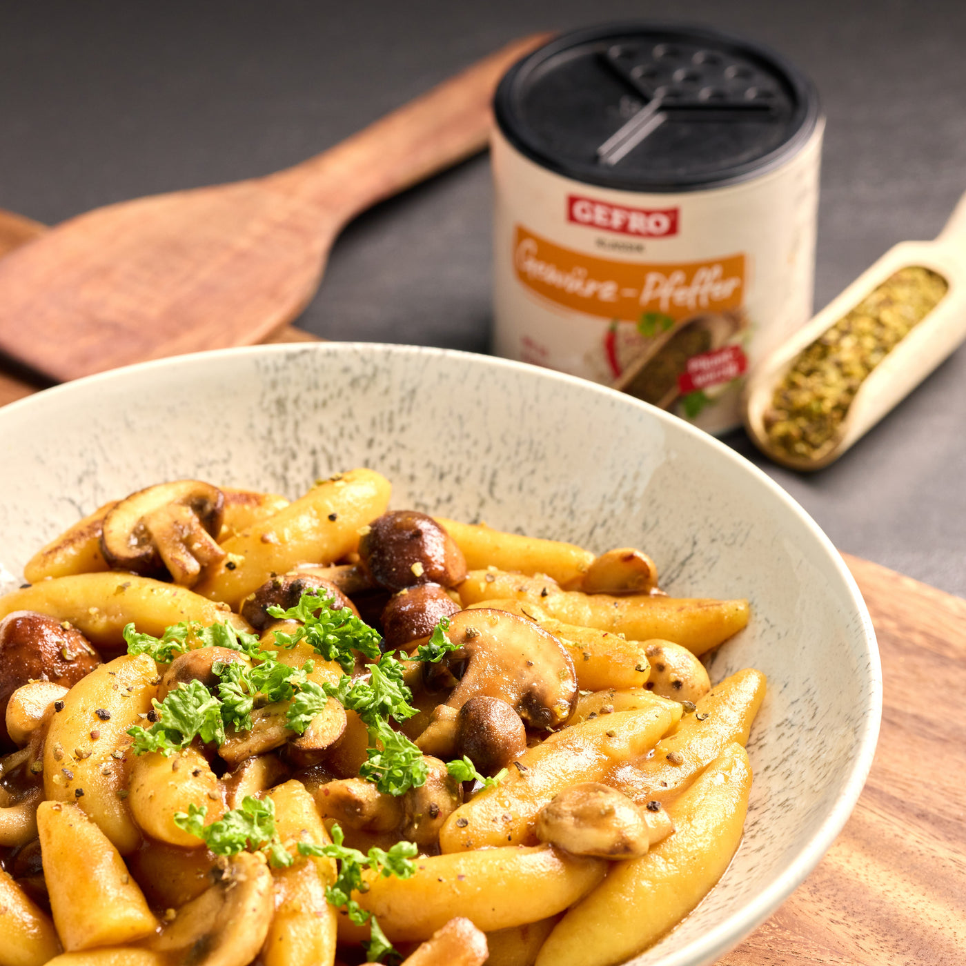 Schupfnudeln mit gebratenen Champignons und Petersilie in einer Schale, daneben Gewürzdose und Holzlöffel