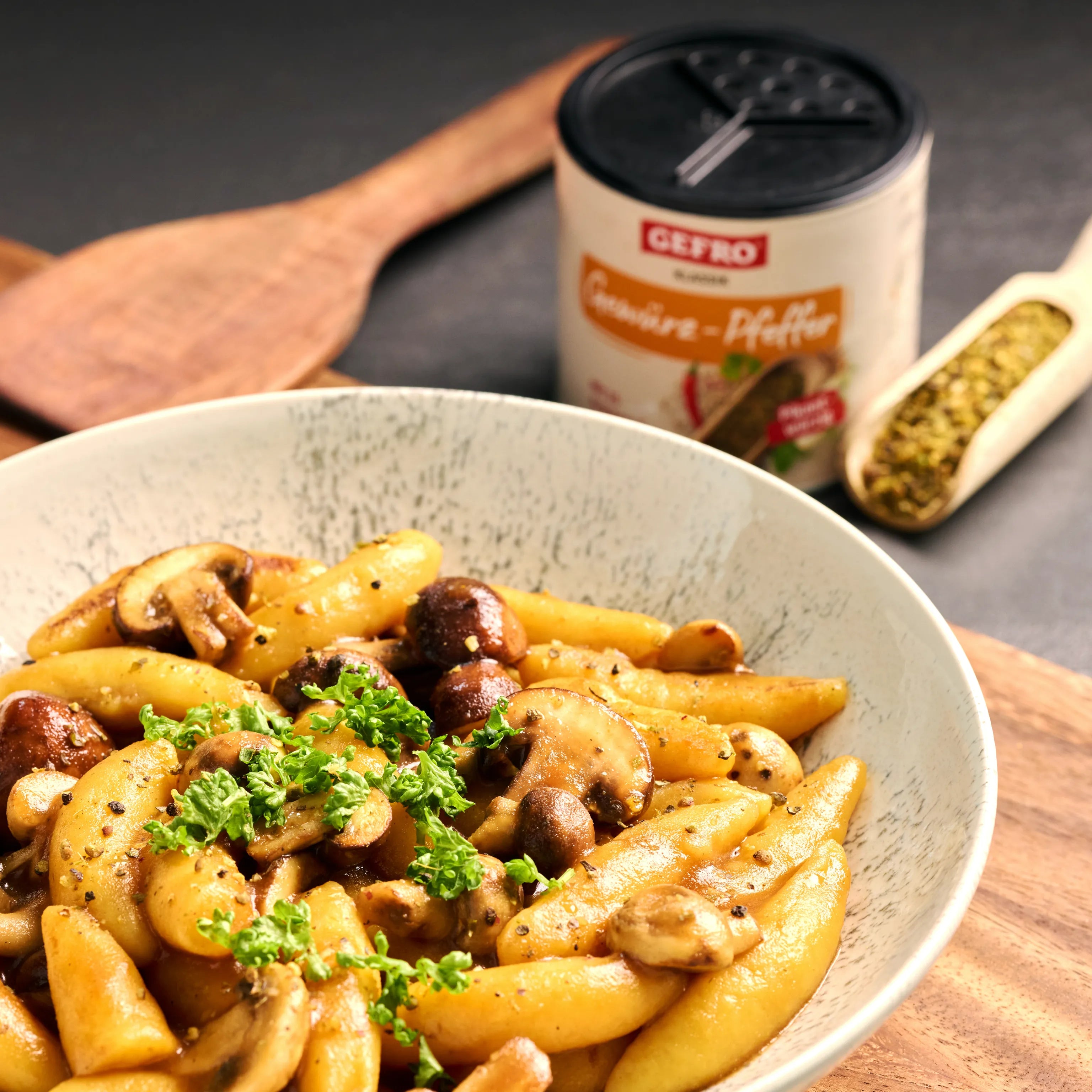 Schupfnudeln mit gebratenen Champignons und Petersilie in einer Schüssel, daneben Gewürzstreuer und Holzlöffel