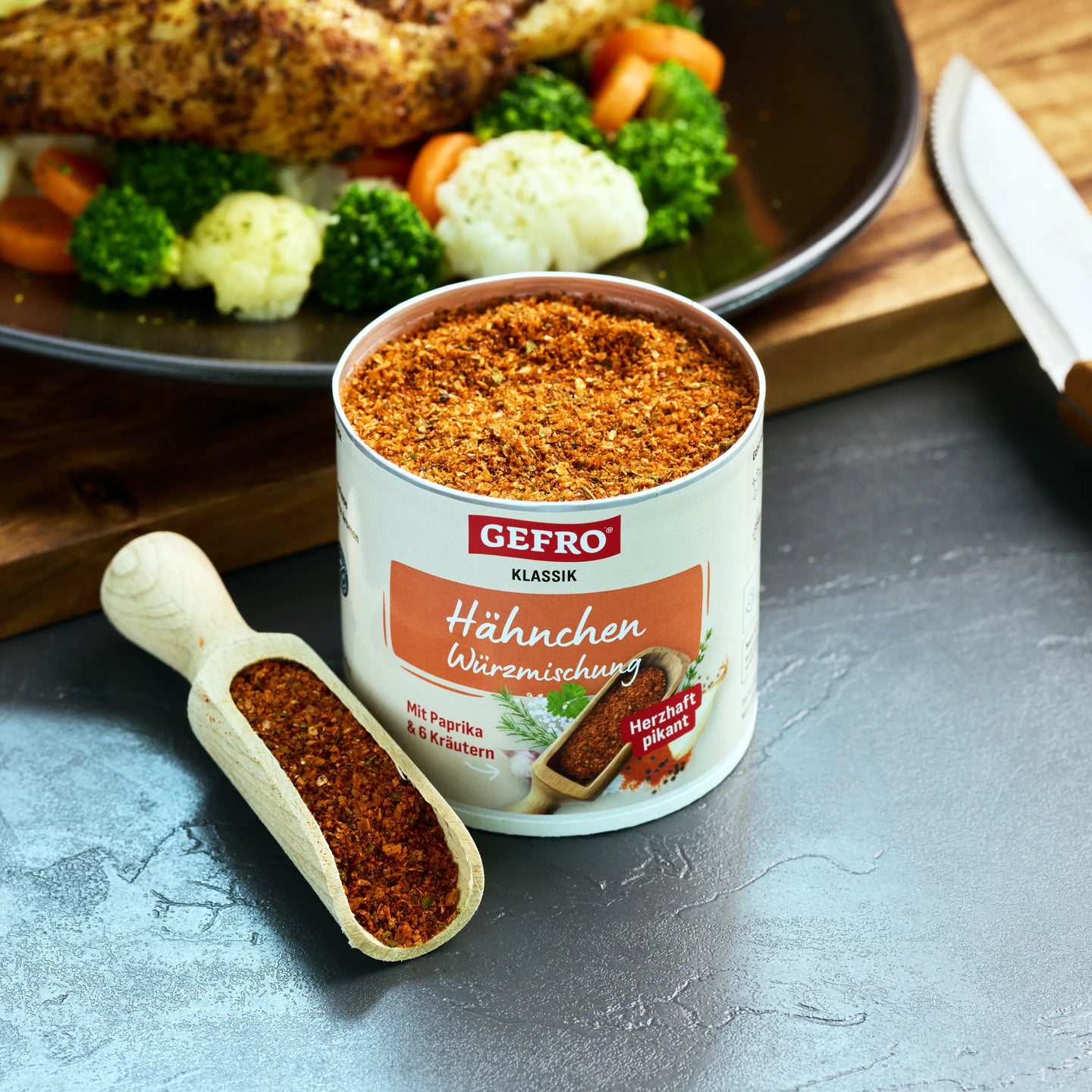 Dose Gefro Hähnchen Würzmischung mit Holzschaufel, dahinter gebratenes Hähnchen mit Brokkoli und Blumenkohl.