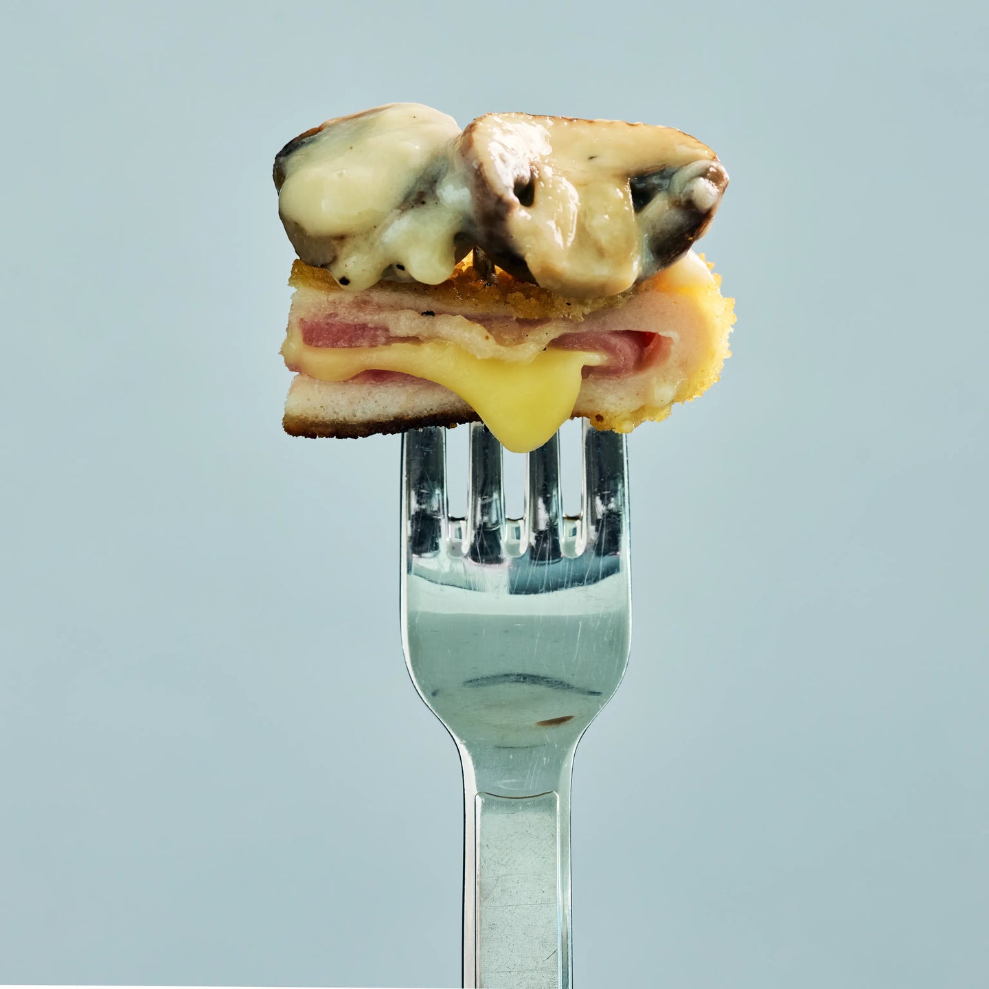 Kleiner Snack mit Schinken und geschmolzenem Käse auf einer Gabel, oben mit zwei überbackenen Champignons belegt