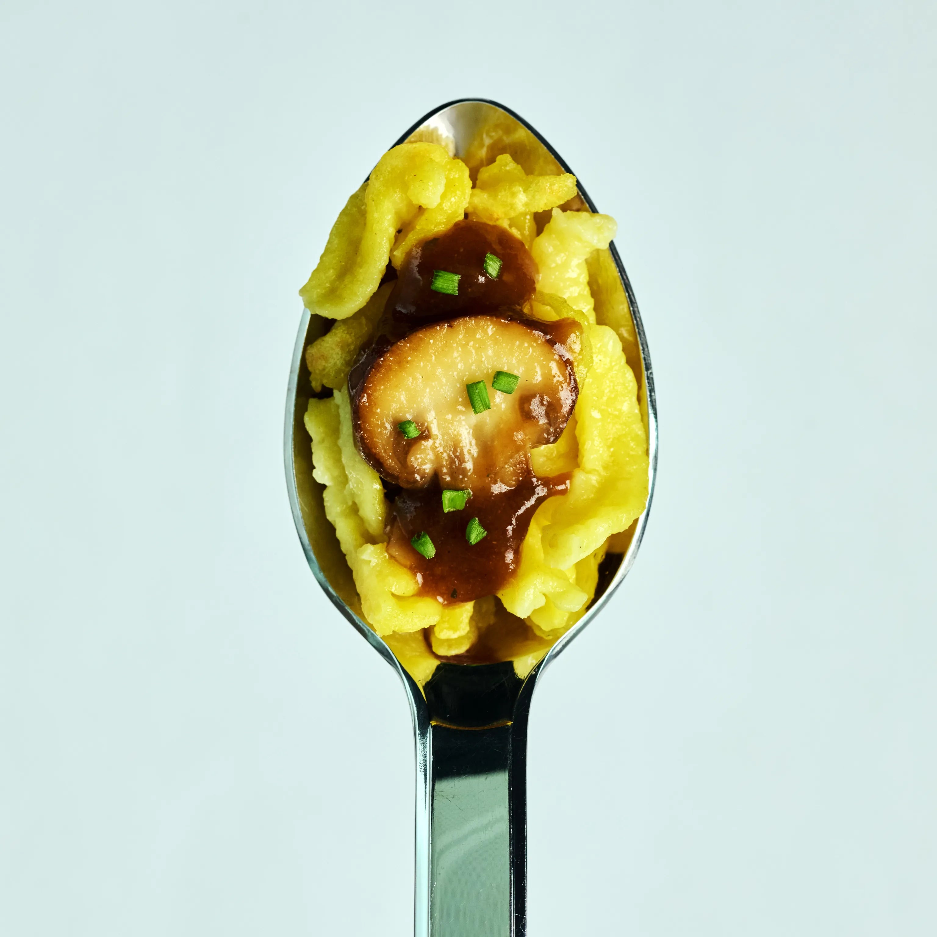 Löffel mit gelben Spätzle, brauner Soße, einer Pilzscheibe und Schnittlauch vor hellem Hintergrund