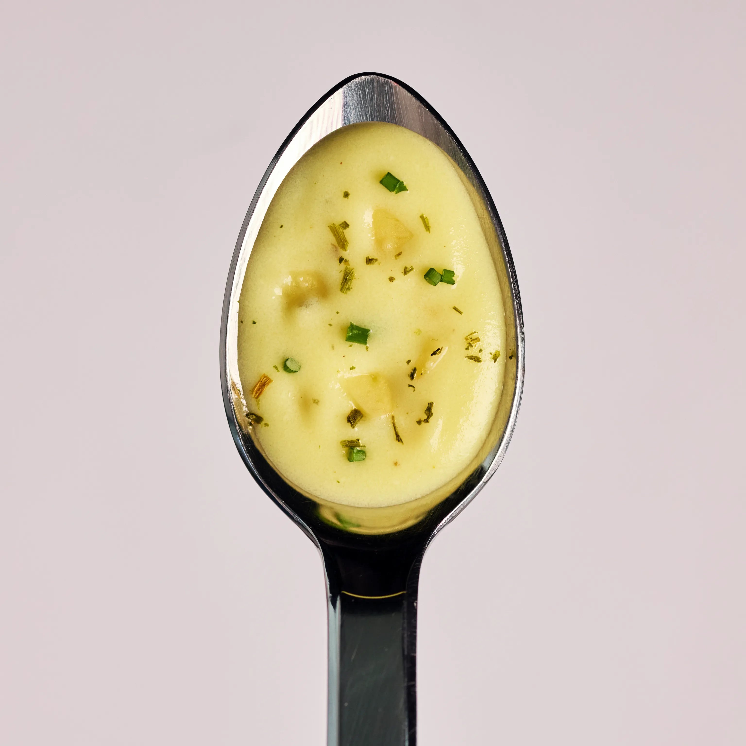 Löffel mit cremiger Kartoffelsuppe und kleinen Kräuterstücken vor hellem Hintergrund, Nahaufnahme des Produkts