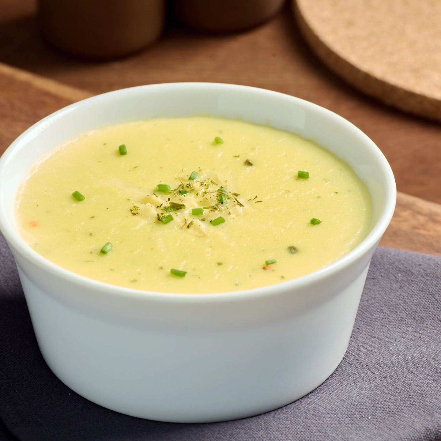 Weiße Schale mit cremiger Kartoffelsuppe, garniert mit Schnittlauch, auf Holzbrett und grauem Tuch serviert