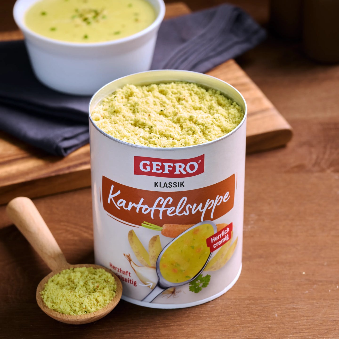 Dose GEFRO Klassik Kartoffelsuppe mit Suppenpulver auf Holztisch, daneben Löffel und eine Schale fertige Suppe.