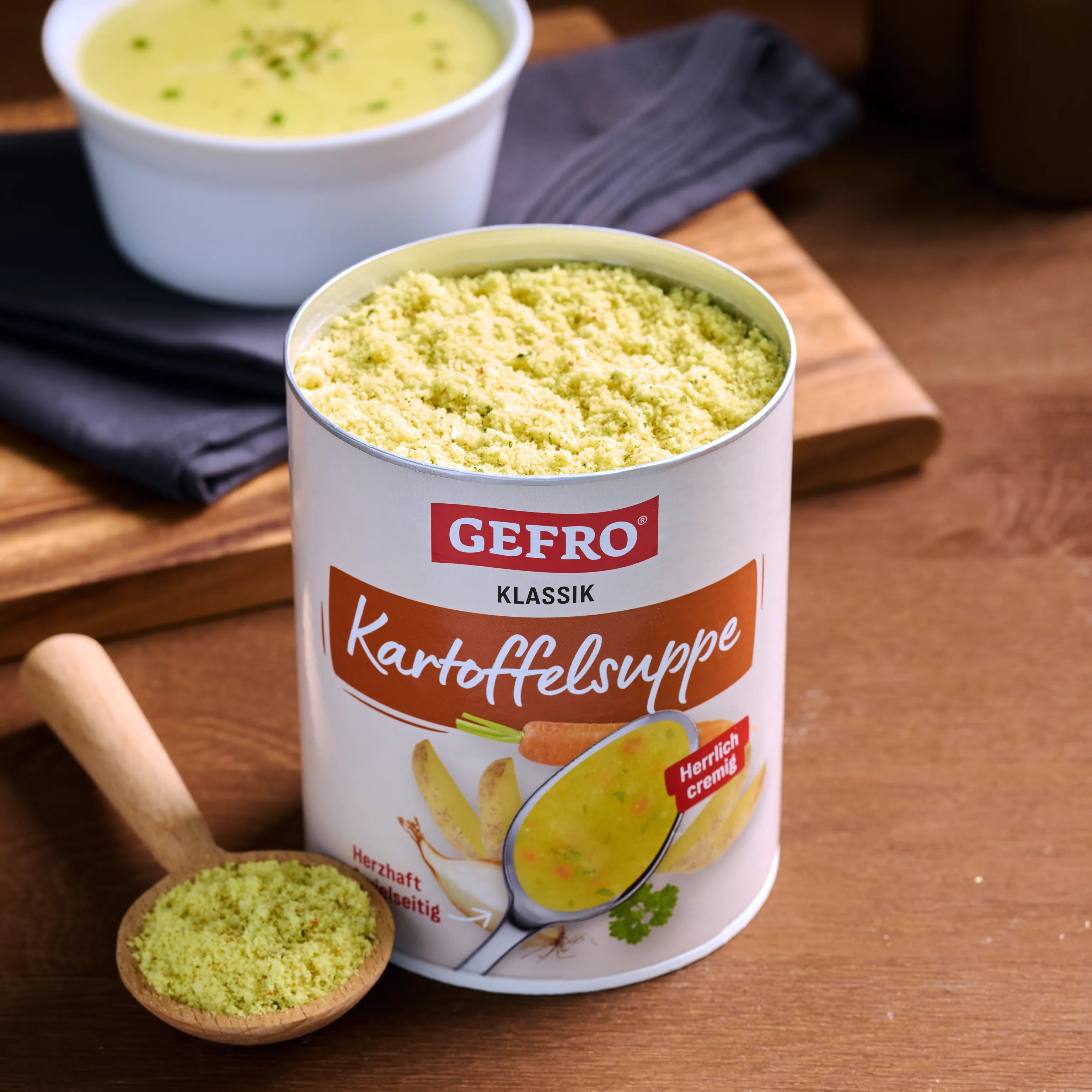 Dose GEFRO Klassik Kartoffelsuppe mit Suppenpulver auf Holztisch, daneben Löffel und eine Schale fertige Suppe.