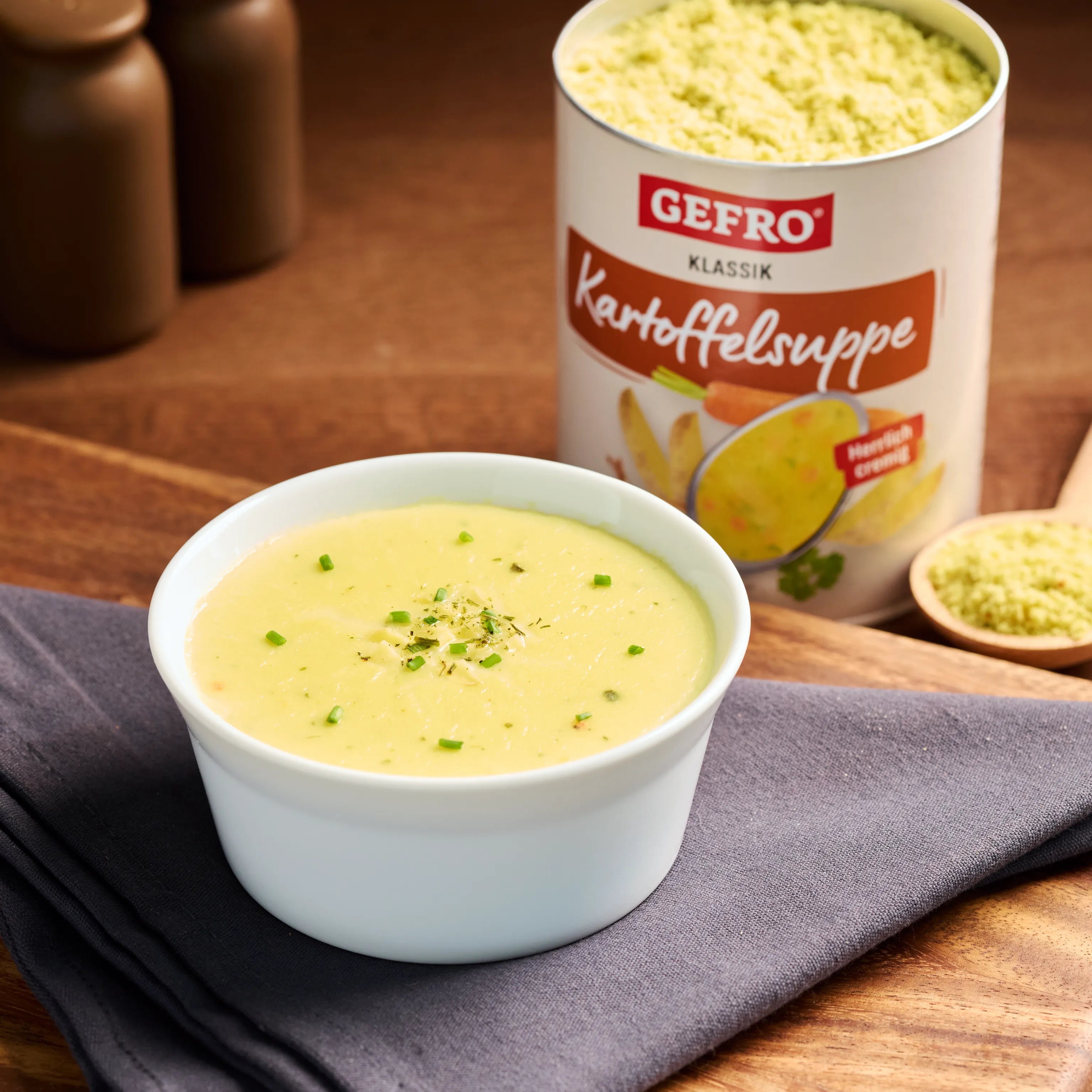 Schale mit cremiger Kartoffelsuppe auf grauem Tuch, daneben Dose Gefro Klassik Kartoffelsuppe auf Holztisch