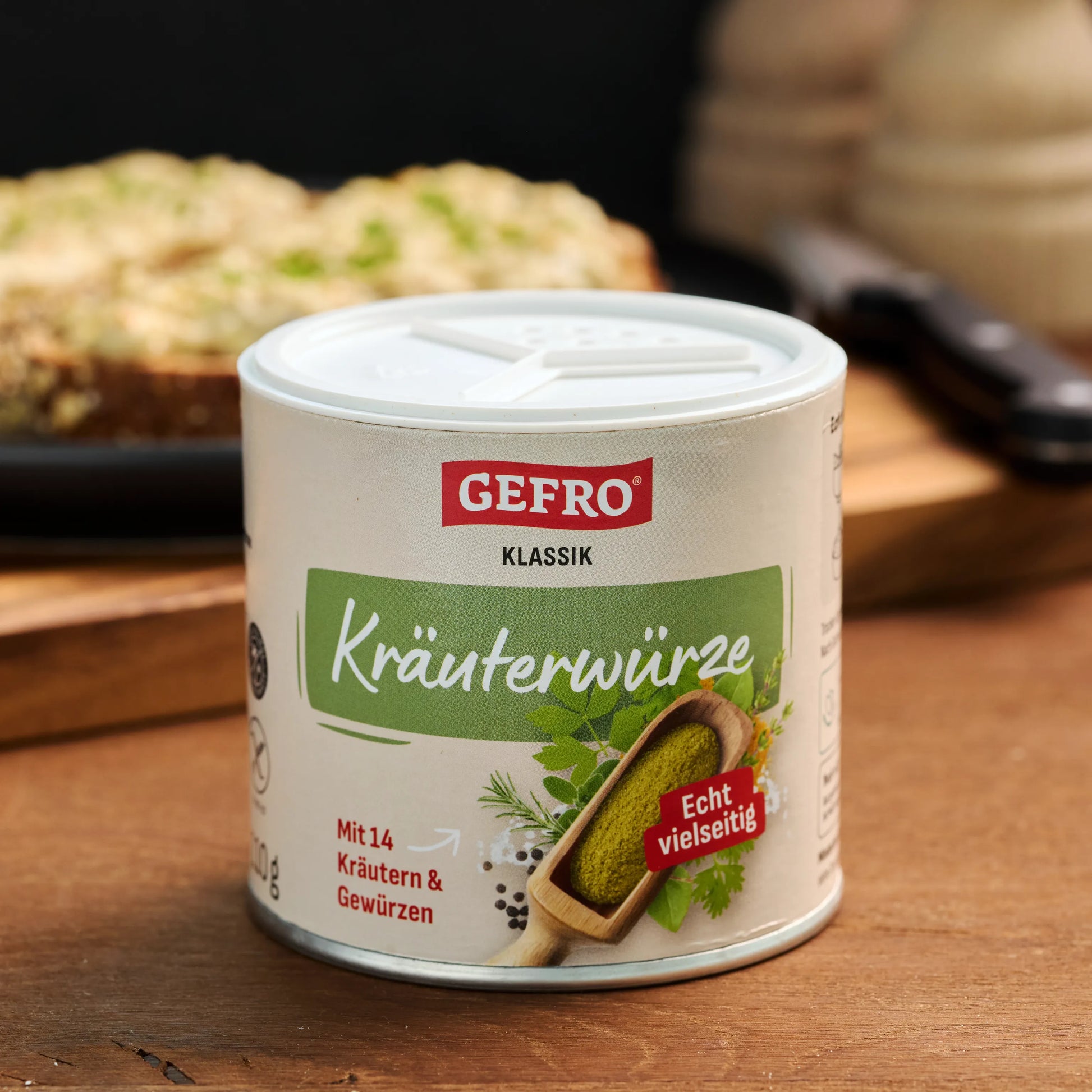 Dose GEFRO Klassik Kräuterwürze auf Holztisch, vorne gezeigt, mit unscharfem Gericht im Hintergrund