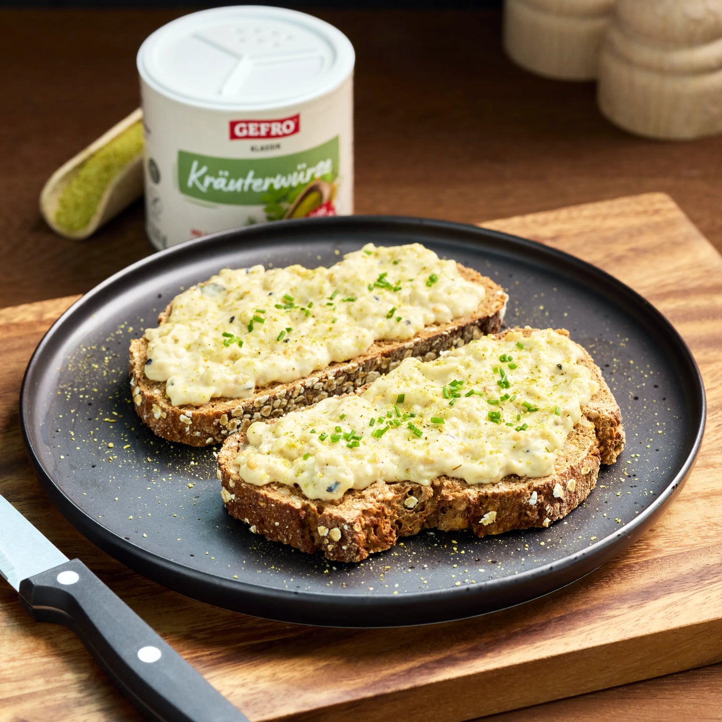Zwei Scheiben Körnerbrot mit cremigem Aufstrich und Schnittlauch auf schwarzem Teller, daneben Kräuterwürze im Hintergrund