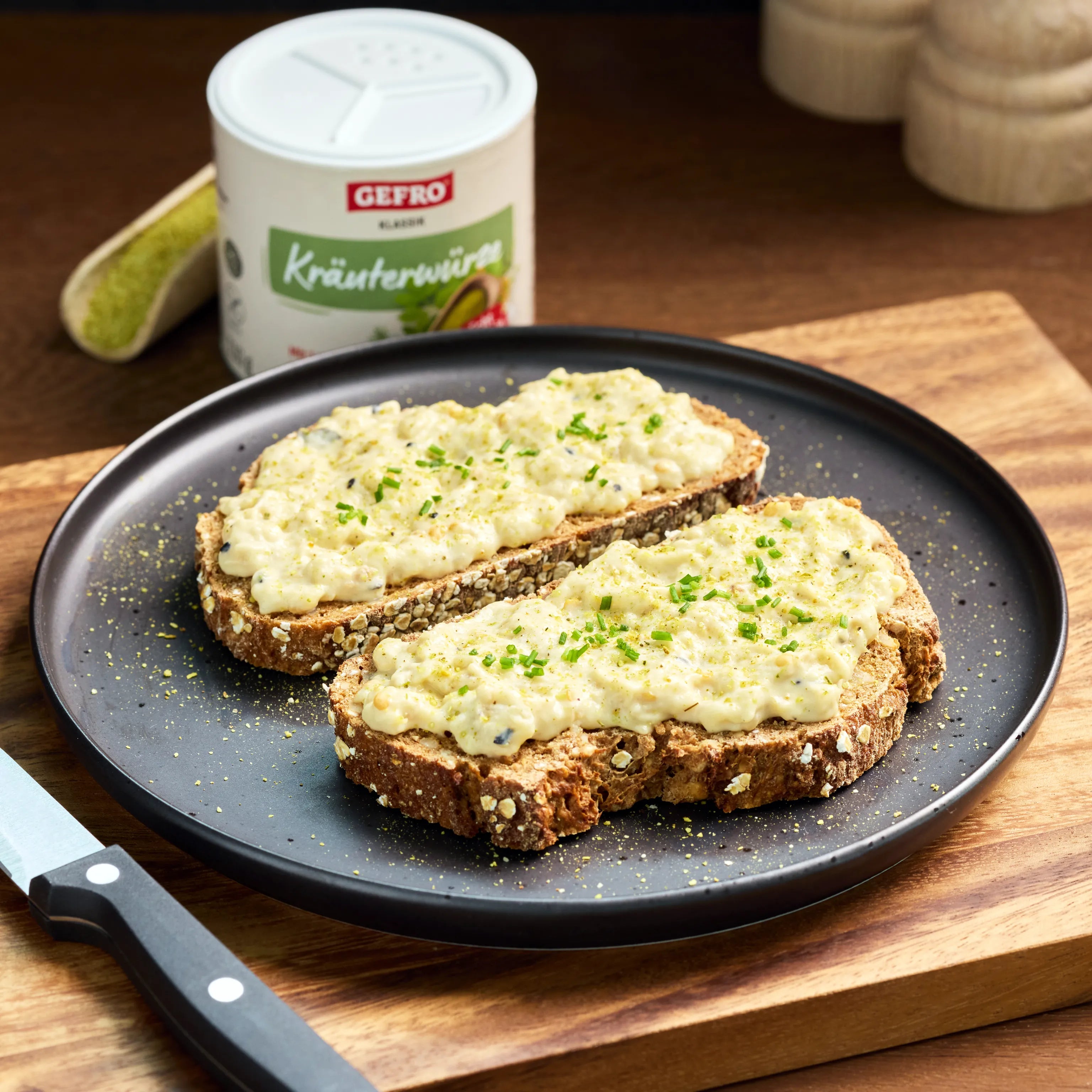 Zwei Scheiben Körnerbrot mit cremigem Aufstrich und Schnittlauch auf schwarzem Teller, daneben Kräuterwürze im Hintergrund