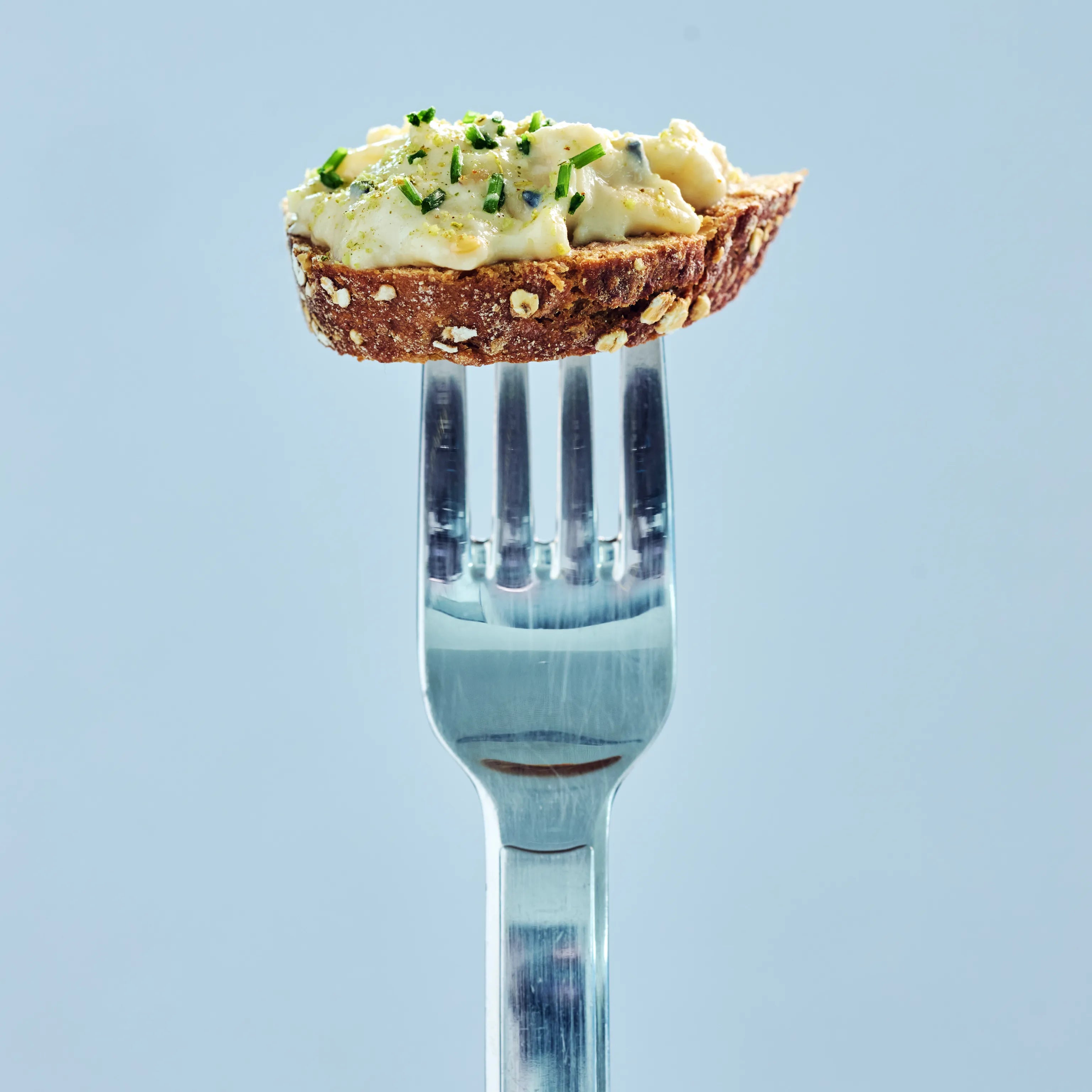 Kleine Scheibe Körnerbrot mit cremigem Aufstrich und Schnittlauch auf einer Gabel vor hellem blauem Hintergrund