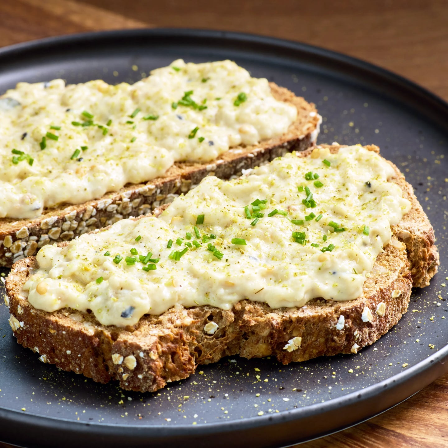 Zwei Scheiben Körnerbrot mit cremigem Eiersalat und Schnittlauch auf einem dunklen Teller serviert.