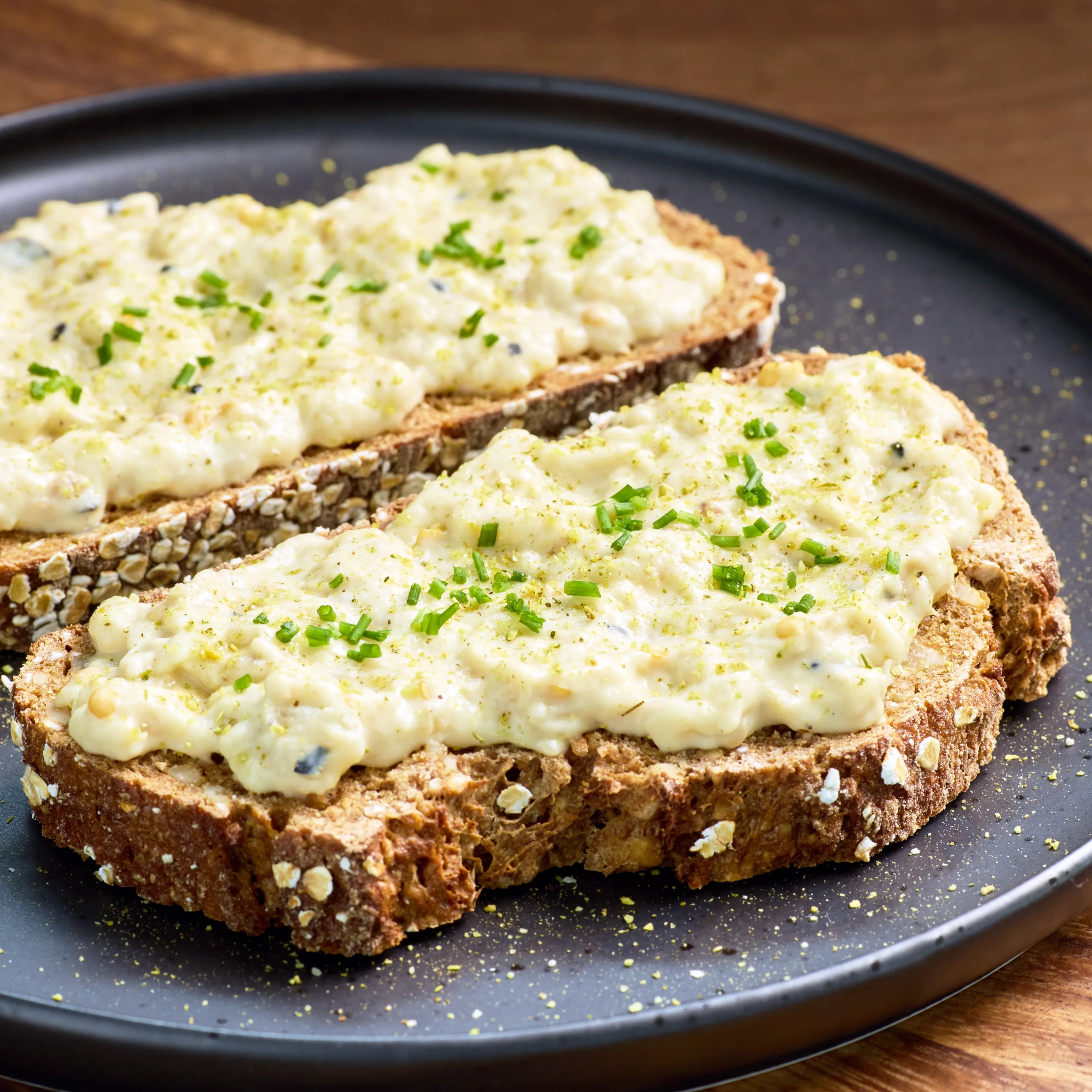 Zwei Scheiben Körnerbrot mit cremigem Eiersalat und Schnittlauch auf einem dunklen Teller serviert.