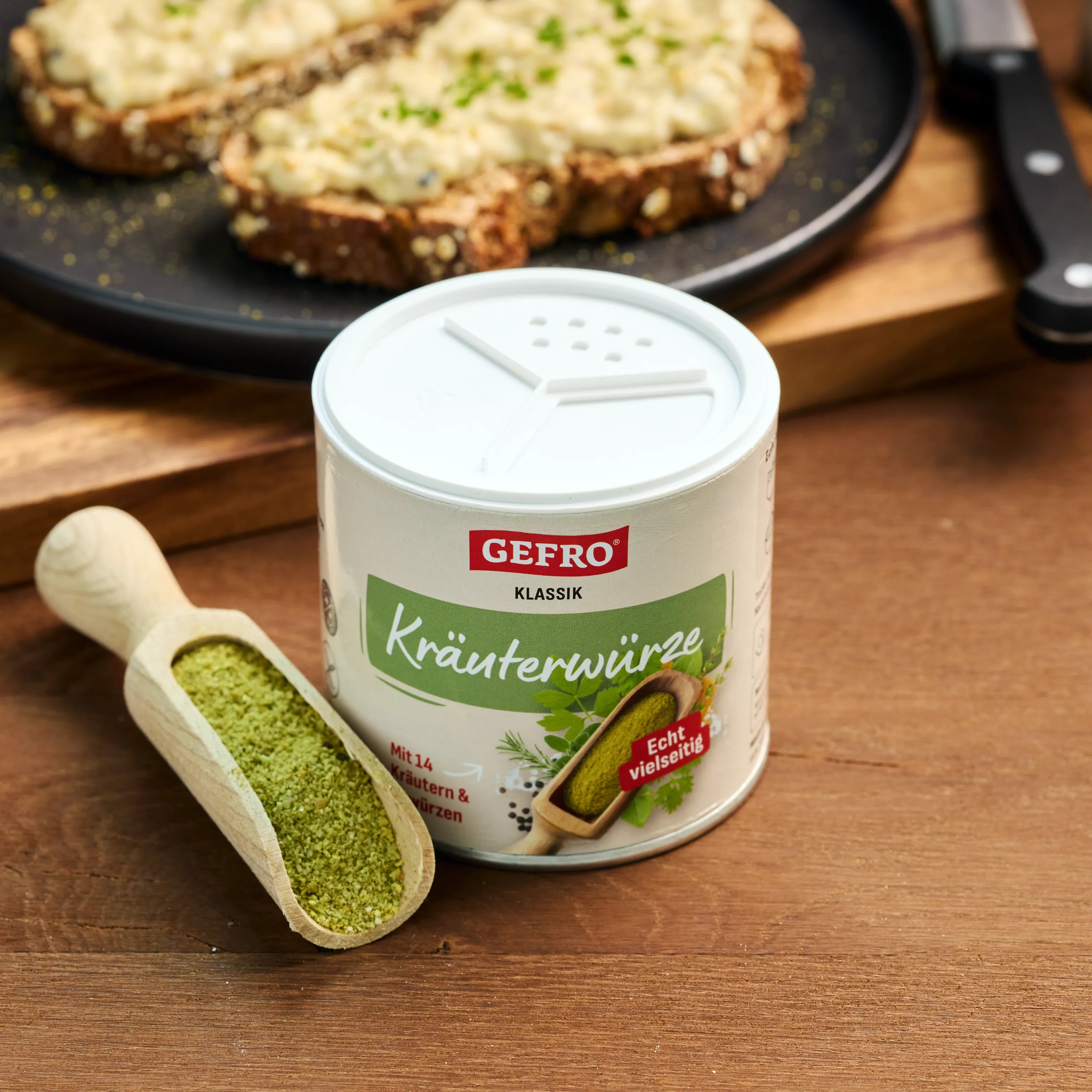 Dose GEFRO Klassik Kräuterwürze mit Holzschaufel auf Tisch, im Hintergrund belegte Brotscheiben.
