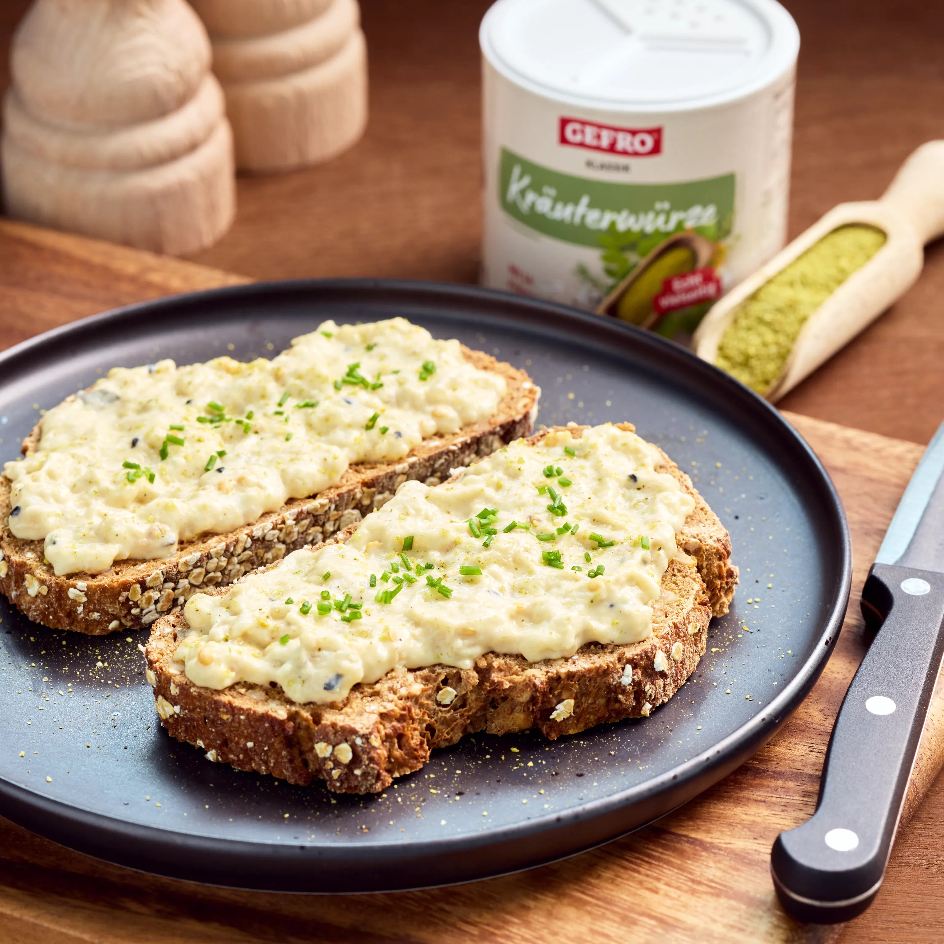 Zwei Brotscheiben mit cremigem Aufstrich und Schnittlauch auf schwarzem Teller, daneben Kräuterwürze und Messer