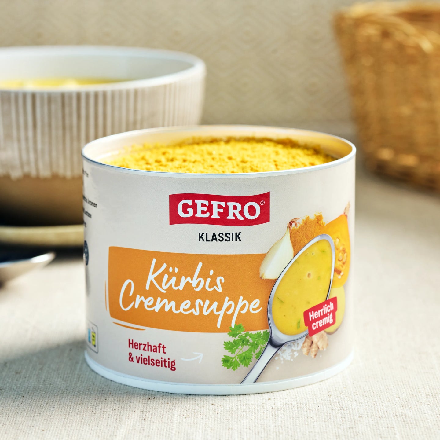 Dose GEFRO Klassik Kürbis Cremesuppe auf Tisch, im Hintergrund eine Schüssel mit zubereiteter Suppe.