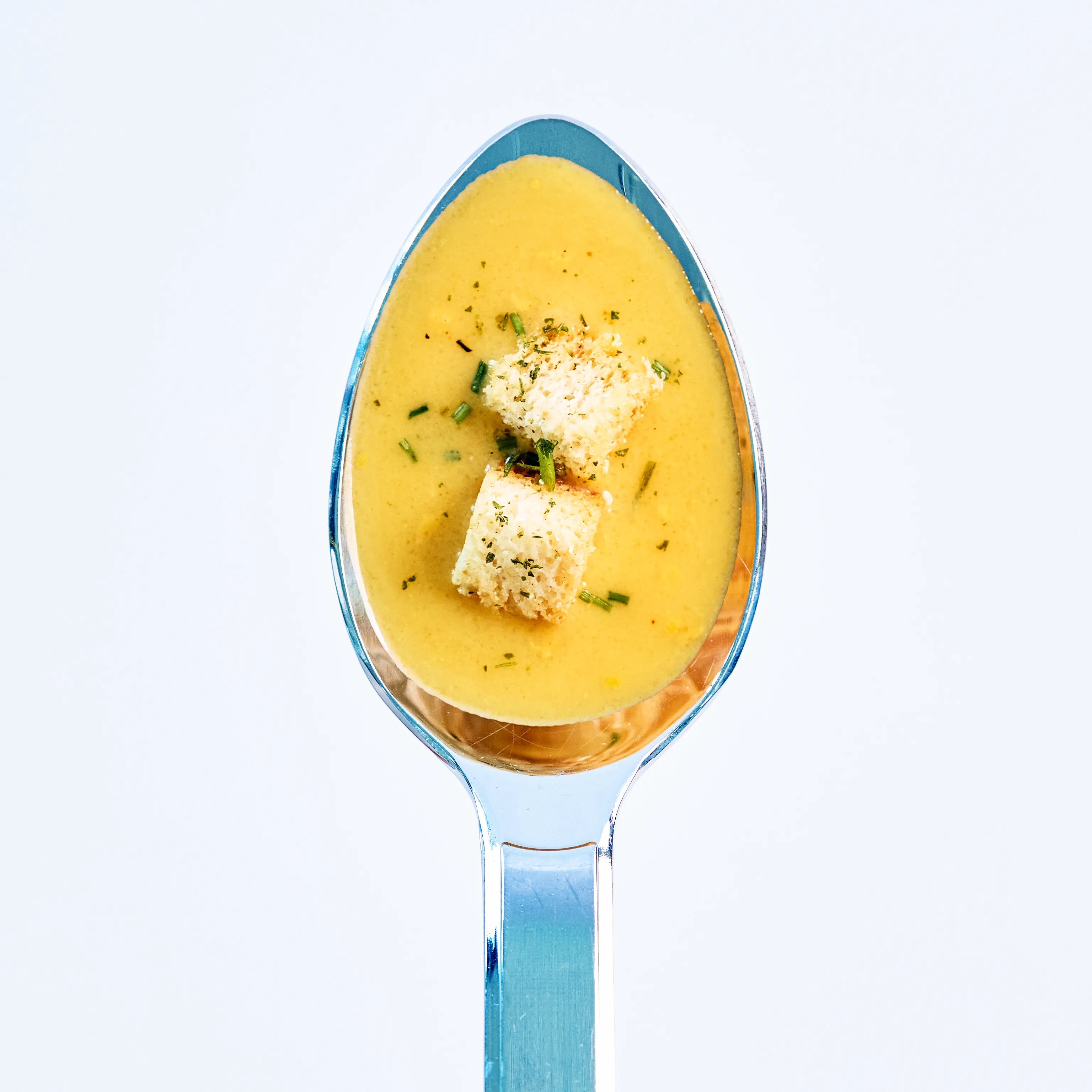 Löffel mit cremiger gelber Suppe, garniert mit zwei Brotwürfeln und frischen Kräutern auf hellem Hintergrund