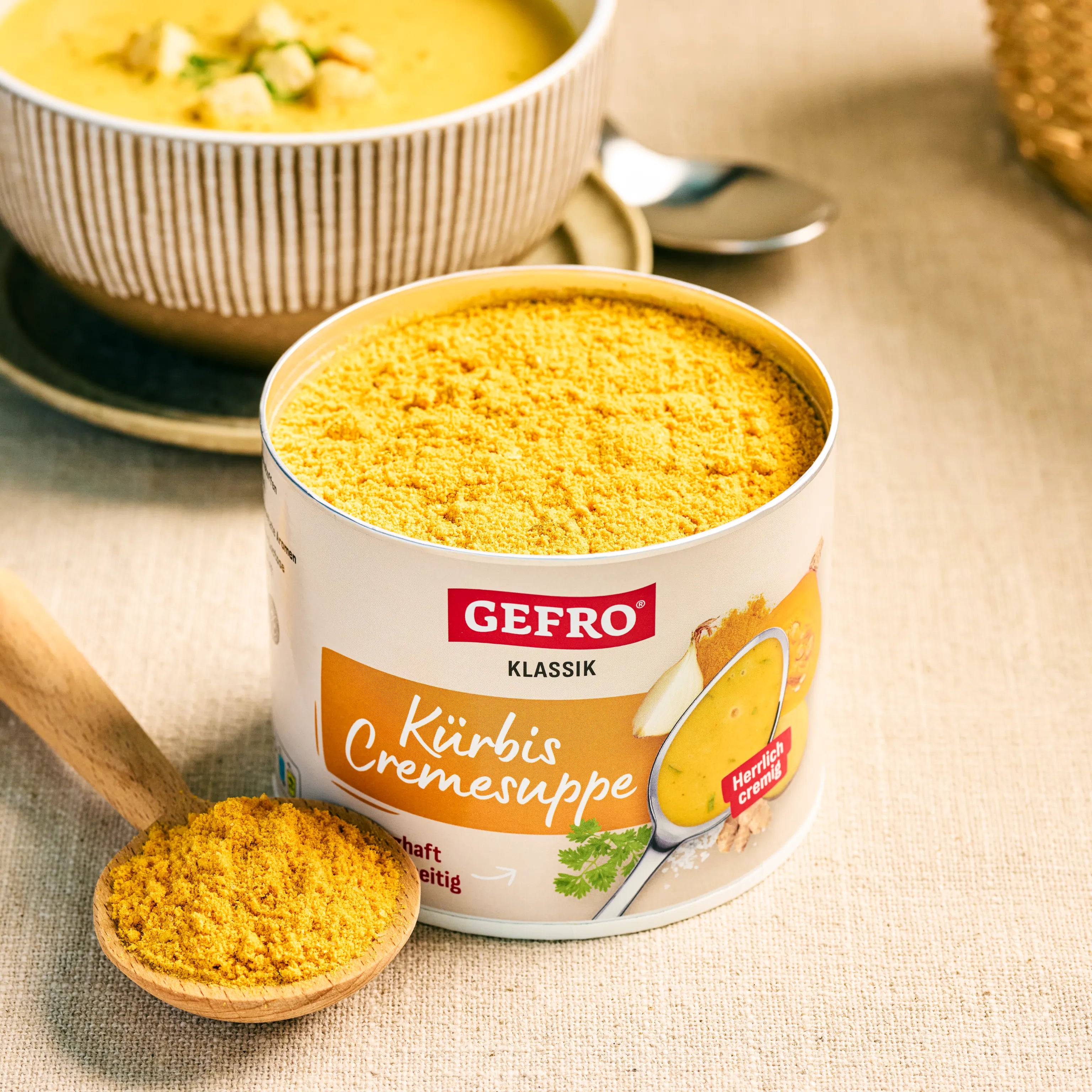 Dose GEFRO Klassik Kürbis Cremesuppe mit Pulver auf Tisch, daneben Holzlöffel und angerichtete Suppe in Schale