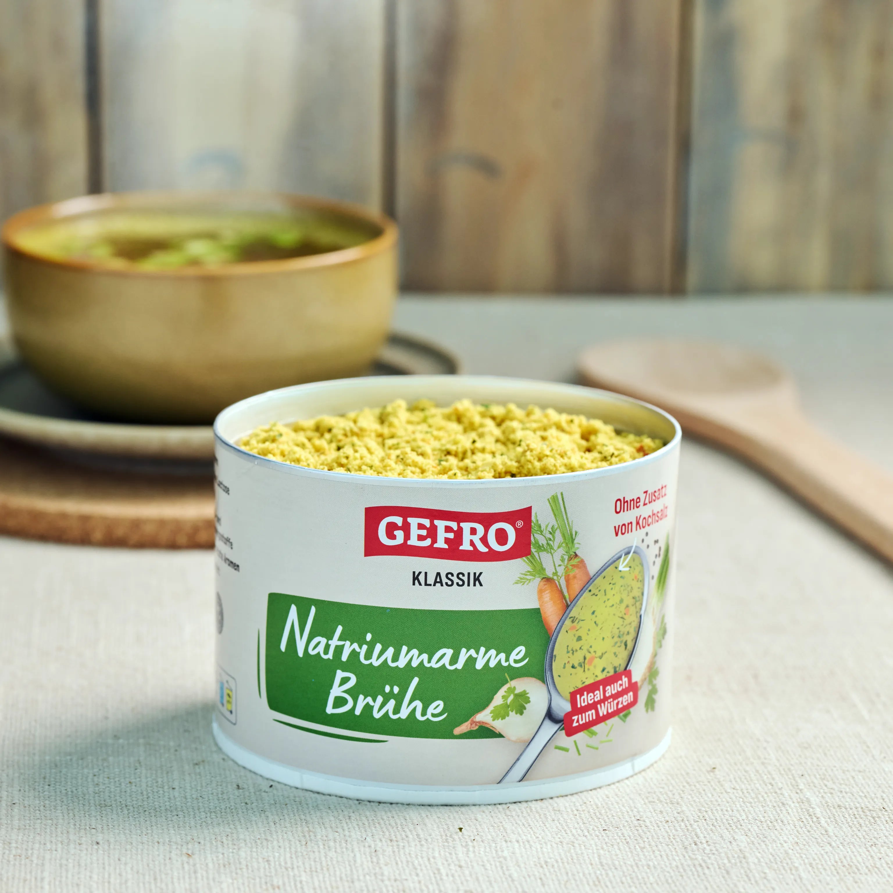 Offene Dose GEFRO Klassik natriumarme Brühe mit gelbem Pulver auf Tisch, im Hintergrund ein Topf mit Suppe.