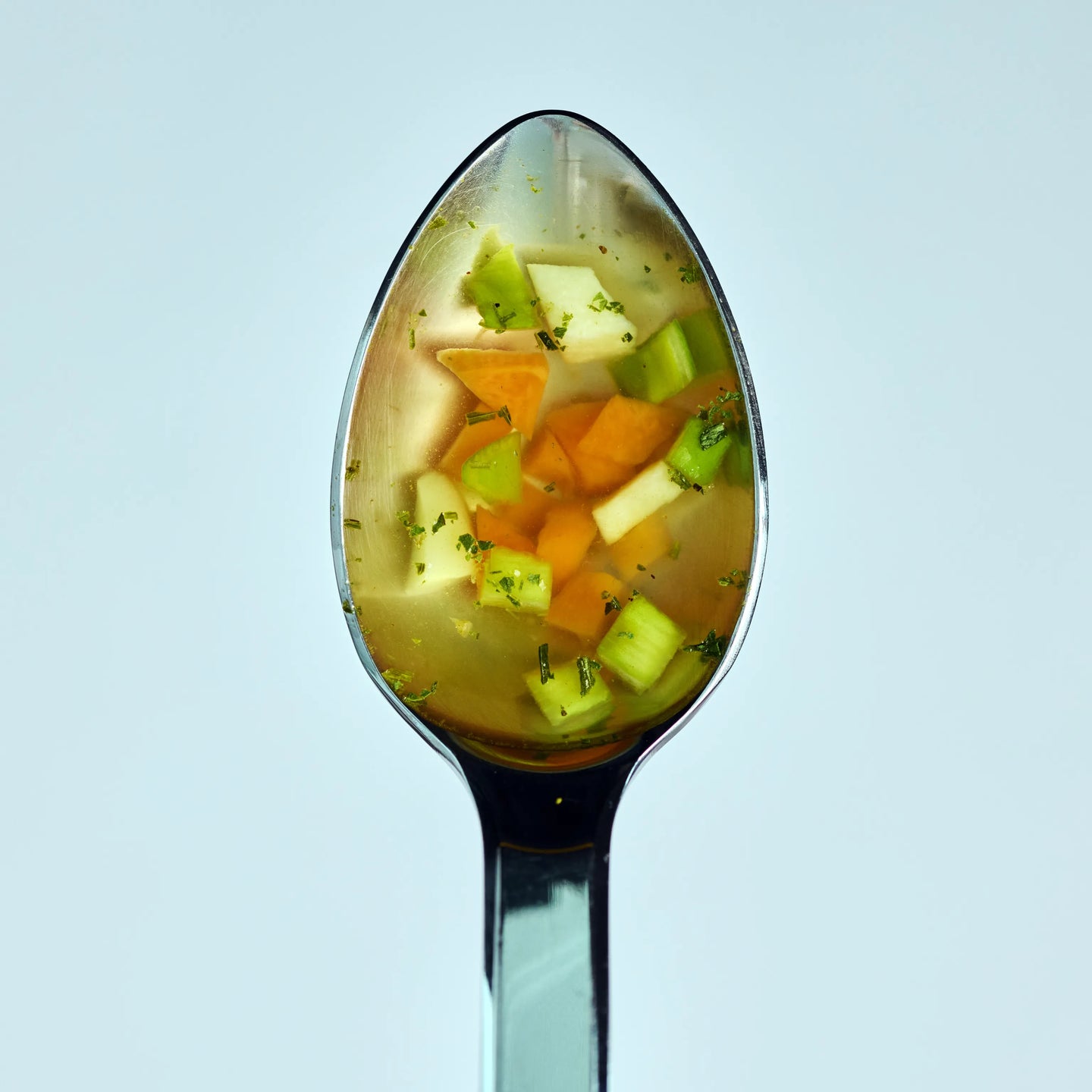 Löffel mit klarer Gemüsesuppe, kleinen Karotten-, Sellerie- und Kartoffelstücken vor hellem Hintergrund