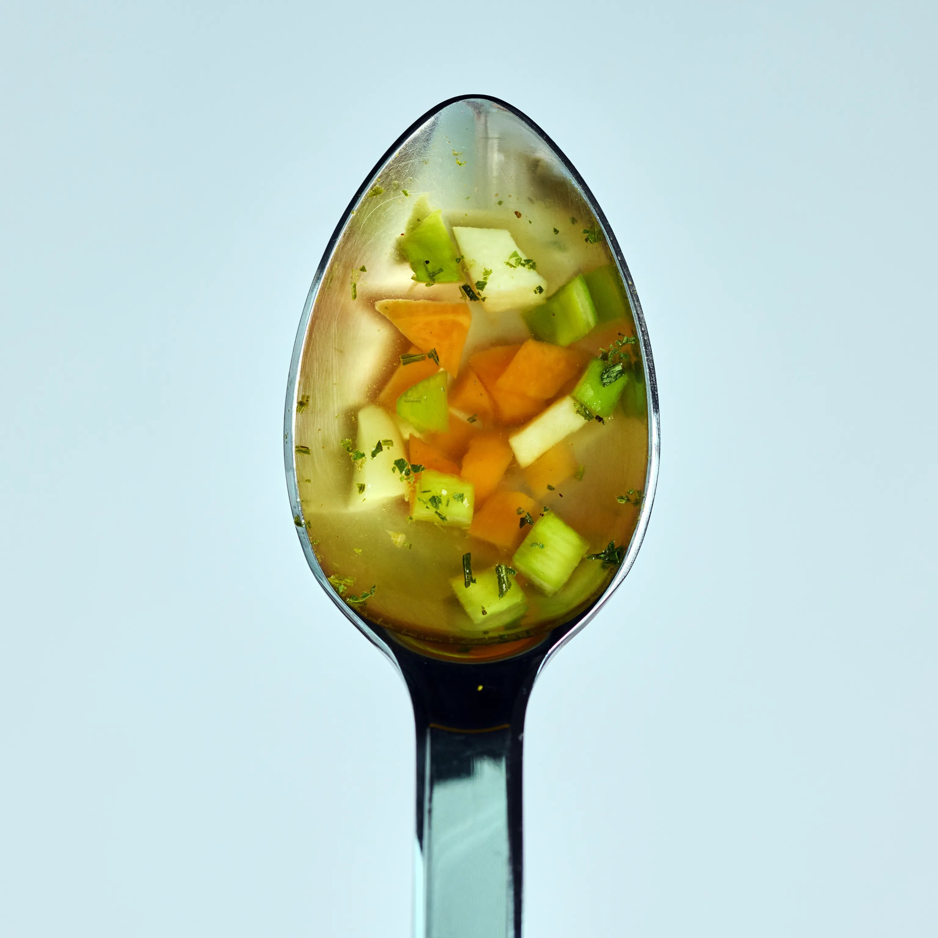 Löffel mit klarer Gemüsesuppe, kleinen Karotten-, Sellerie- und Kartoffelstücken vor hellem Hintergrund