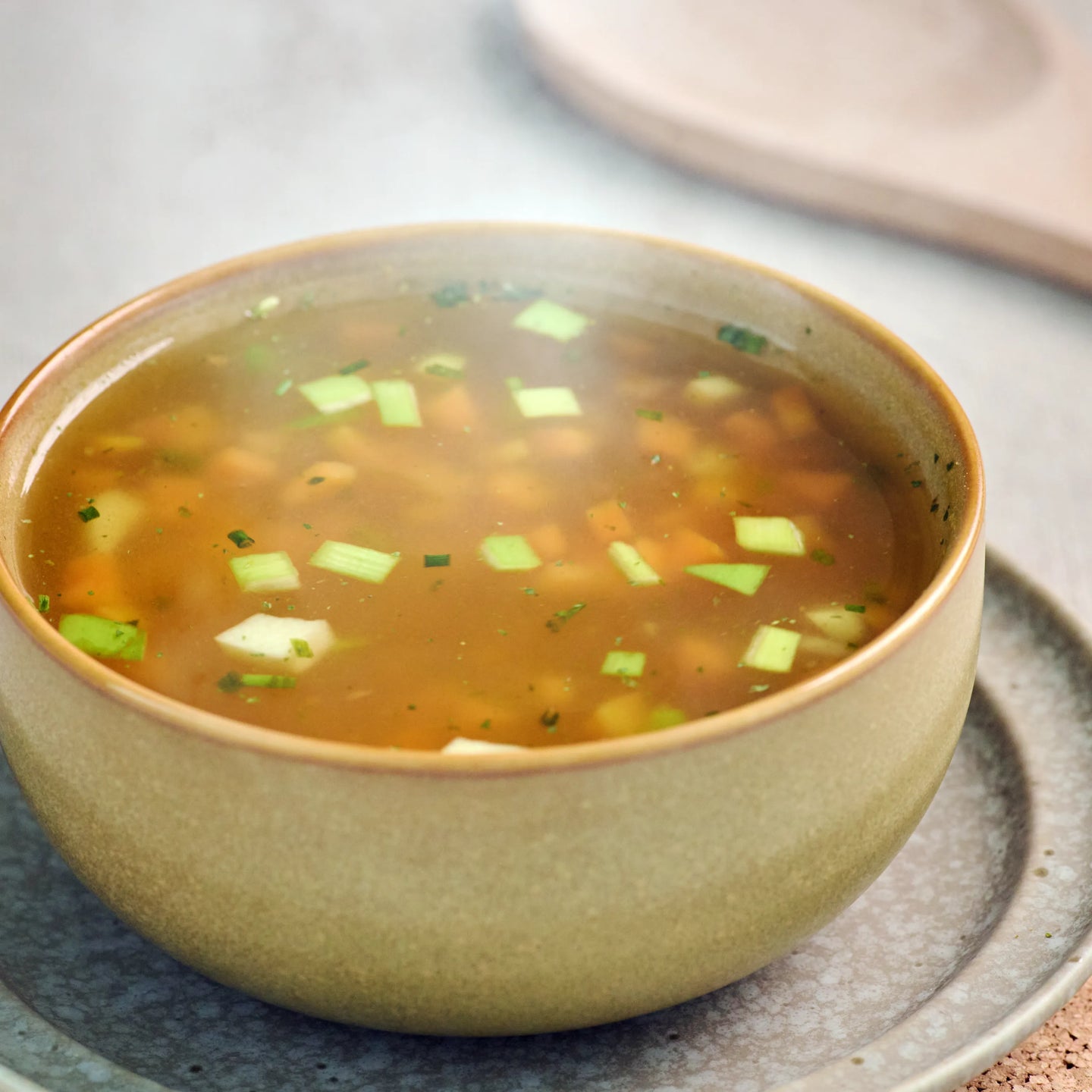 Schale mit klarer Gemüsesuppe auf einem Teller, mit kleinen Gemüsestücken und Frühlingszwiebeln garniert.