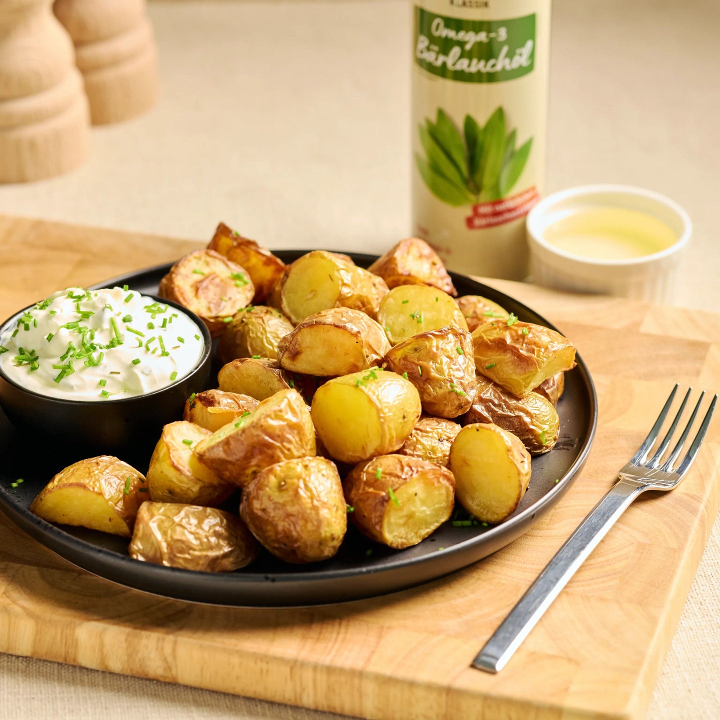 Teller mit knusprigen Ofenkartoffeln und Kräuterquark, daneben eine Flasche Omega-3 Bratöl auf Holzbrett