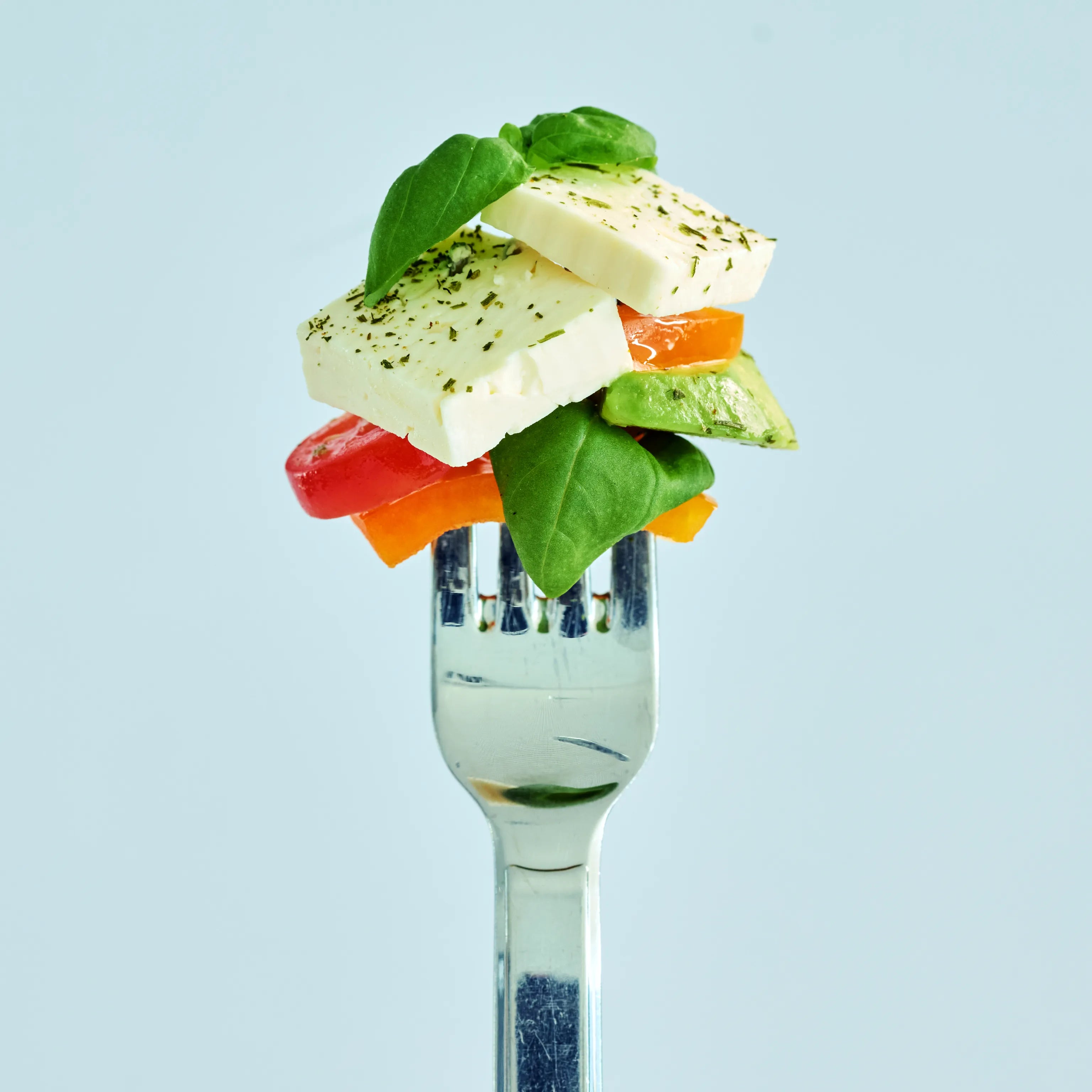 Gabel mit weißen Käsewürfeln, Basilikumblättern, Avocado und buntem Gemüse vor hellem Hintergrund