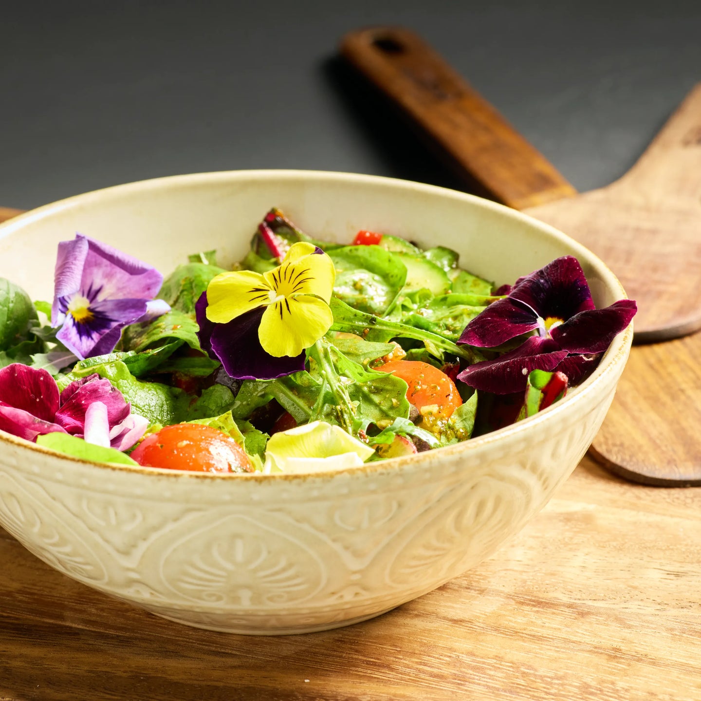 Keramikschüssel mit gemischtem Salat, Tomaten, Gurken und essbaren Blüten auf einem Holztisch.