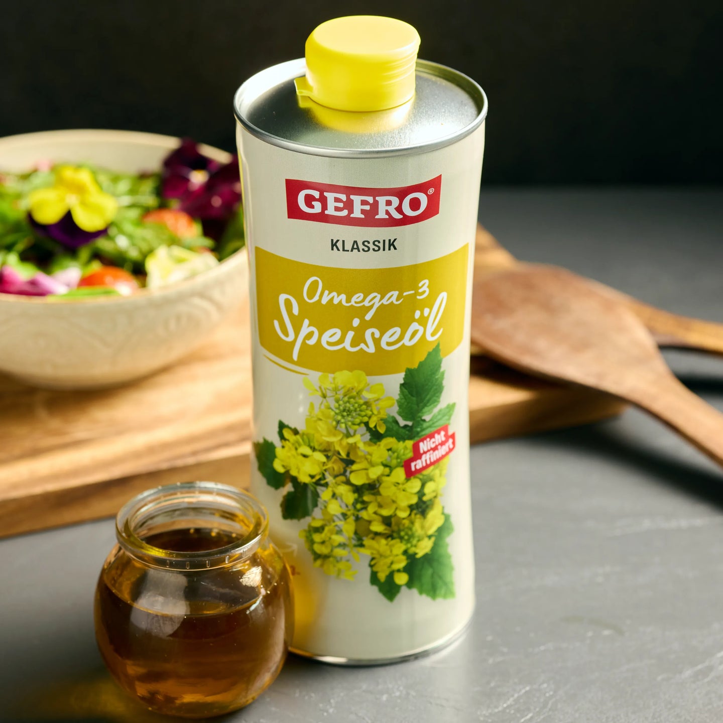 Dose GEFRO Klassik Omega-3 Speiseöl mit gelbem Deckel, davor ein Glas Öl, im Hintergrund Salat und Holzbrett