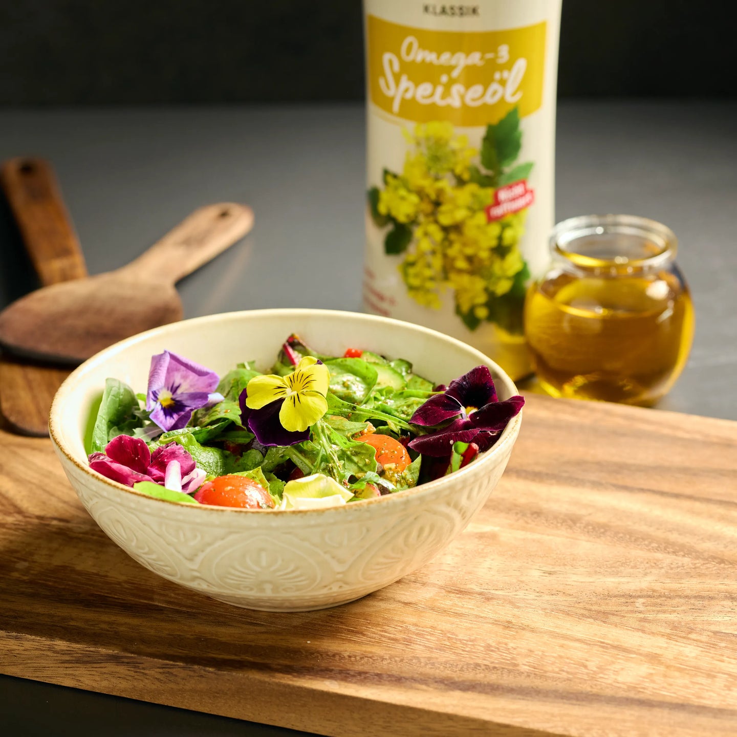 Schale mit frischem Salat und essbaren Blüten auf Holzbrett, daneben Omega-3-Speiseöl und kleines Glas Öl