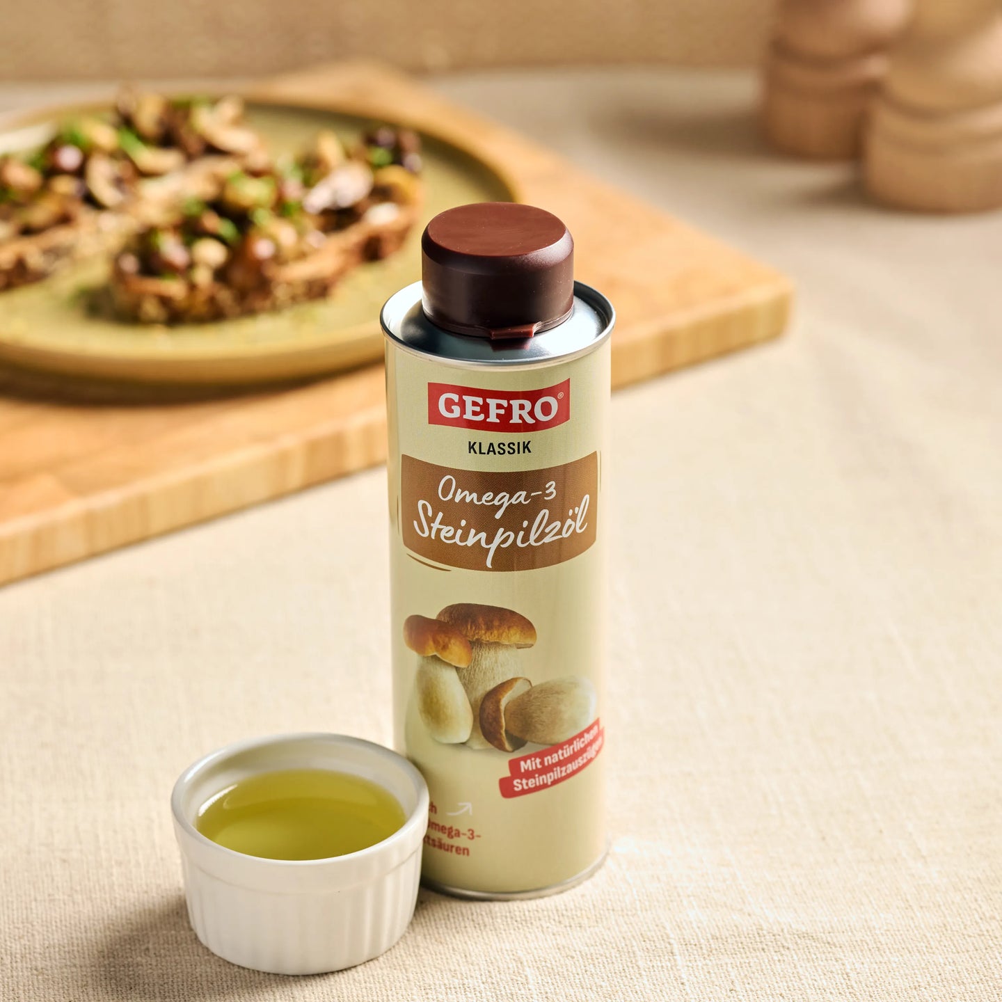 Dose GEFRO Klassik Omega-3 Steinpilzöl mit Schälchen Öl auf dem Tisch, im Hintergrund ein angerichteter Teller.
