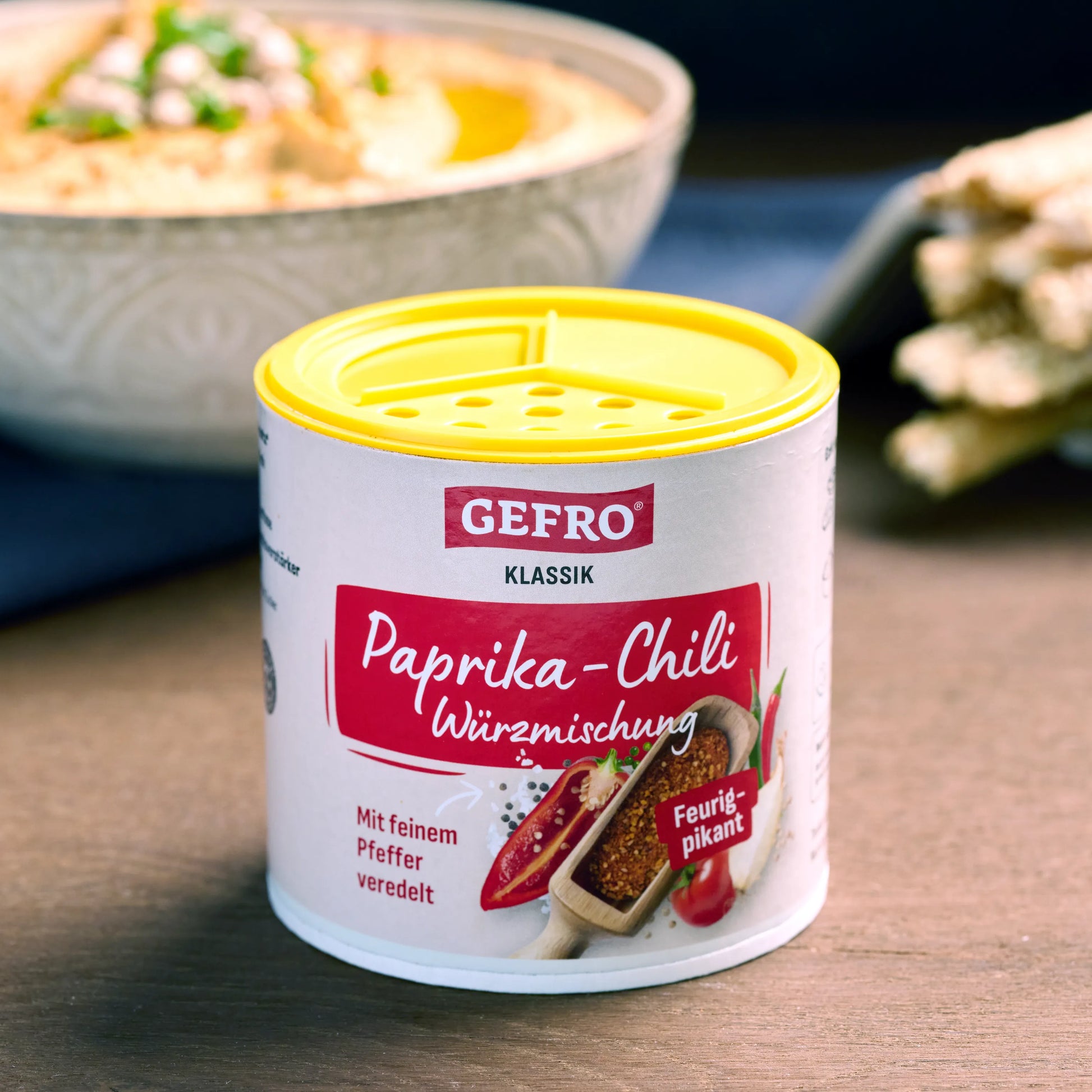 Dose GEFRO Paprika-Chili Würzmischung mit gelbem Streudeckel, davor platziert, im Hintergrund unscharfe Speisen