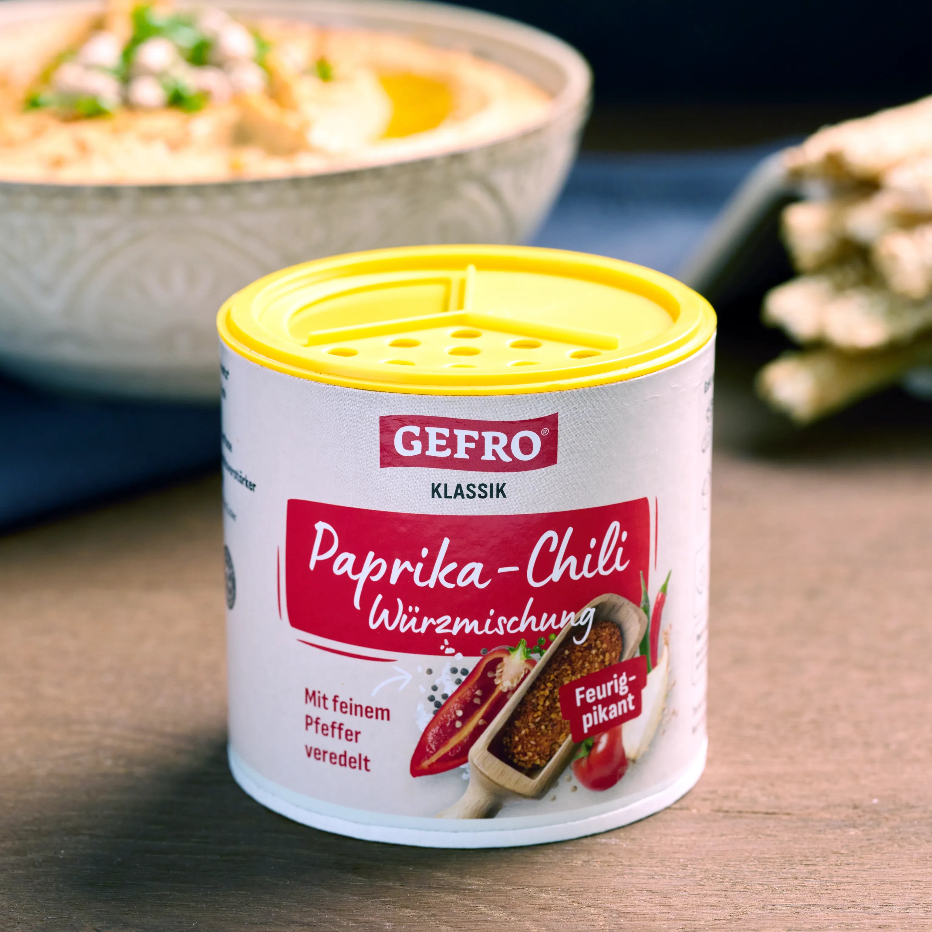 Dose GEFRO Paprika-Chili Würzmischung mit gelbem Streudeckel, davor platziert, im Hintergrund unscharfe Speisen
