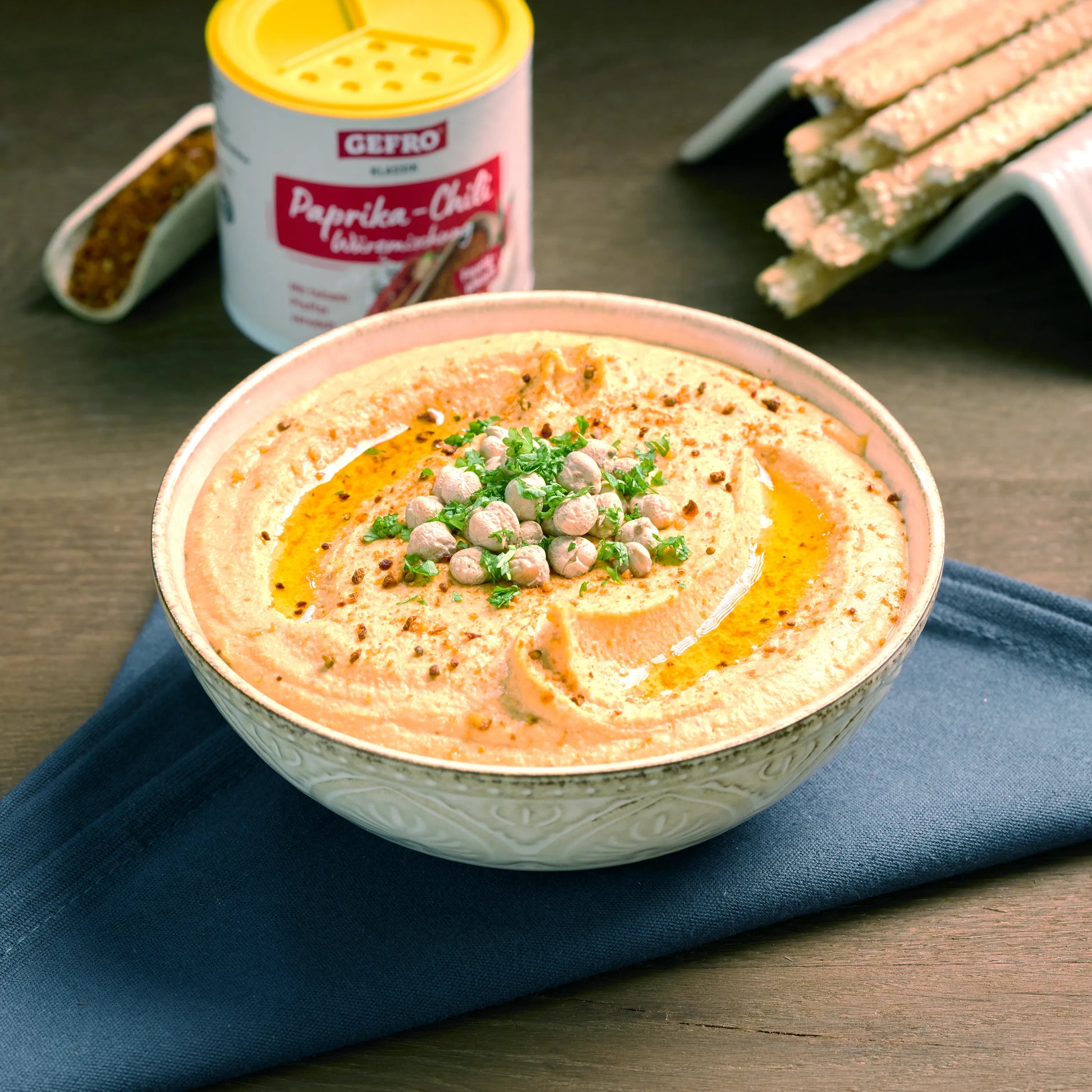 Schale mit cremigem Hummus, garniert mit Kichererbsen, Petersilie und Öl, daneben Gewürz und Grissini.