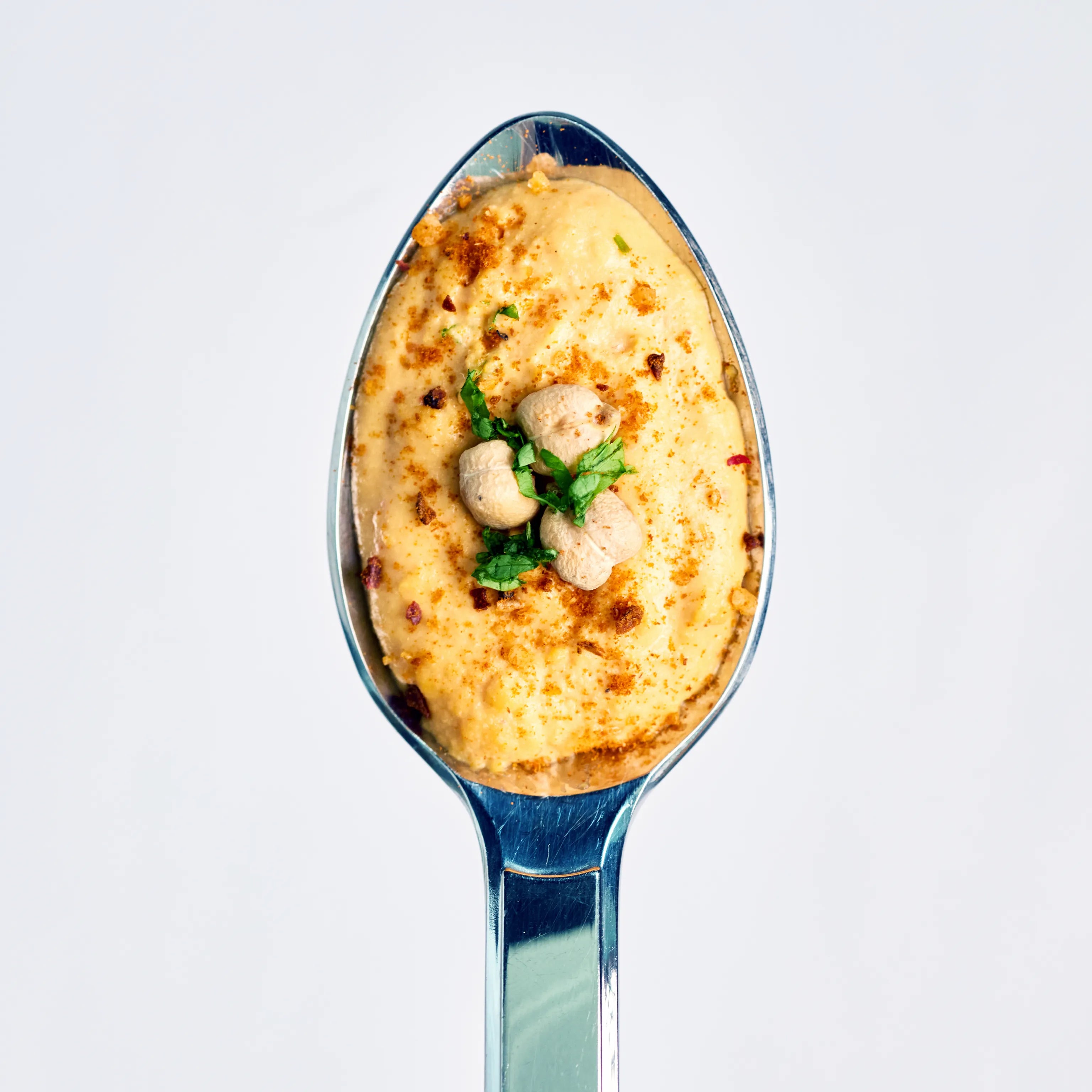 Löffel mit cremigem Hummus, garniert mit Kichererbsen, Kräutern und Gewürzen vor hellem Hintergrund