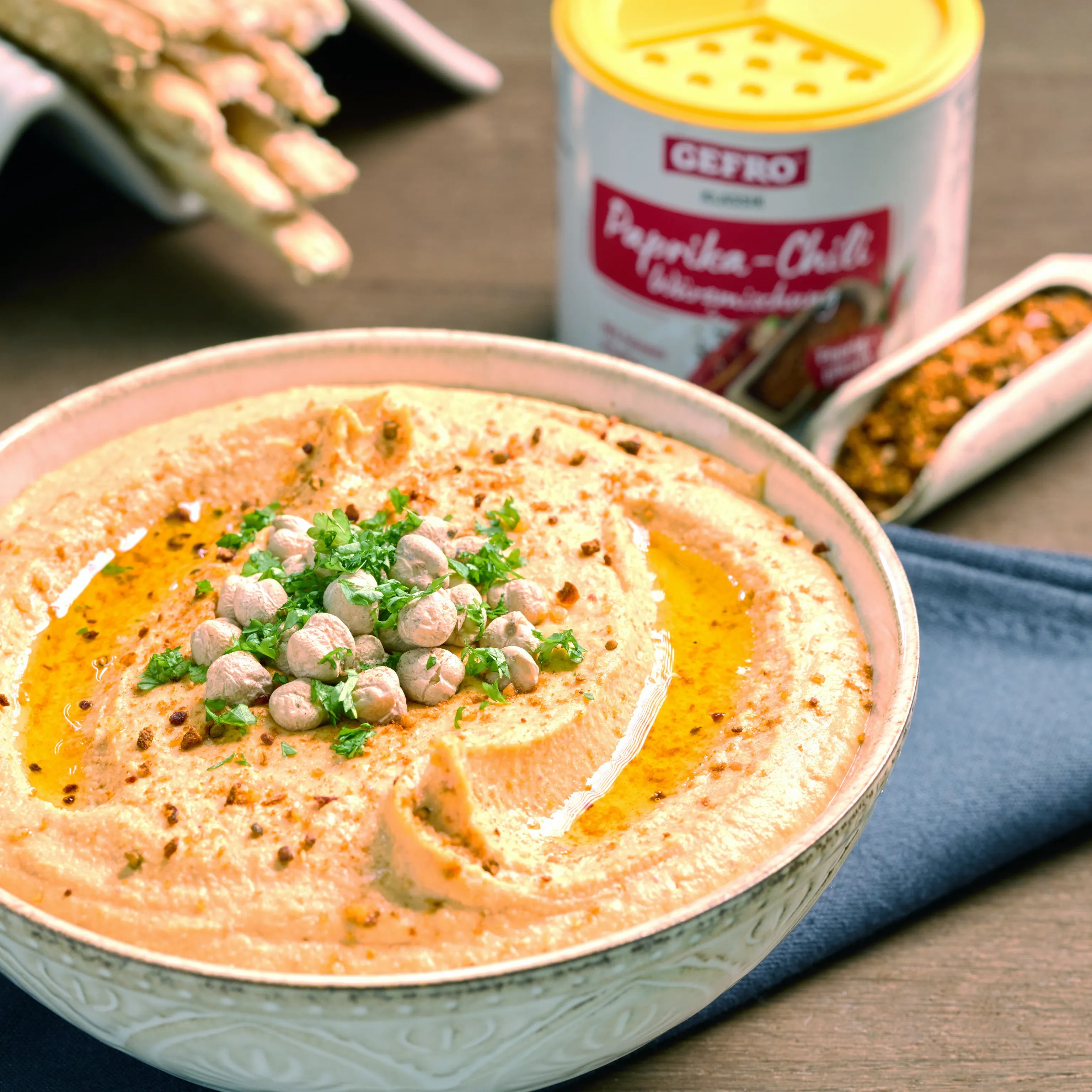 Schale mit cremigem Hummus, garniert mit Kichererbsen, Petersilie und Öl; im Hintergrund Paprika-Chili-Gewürz