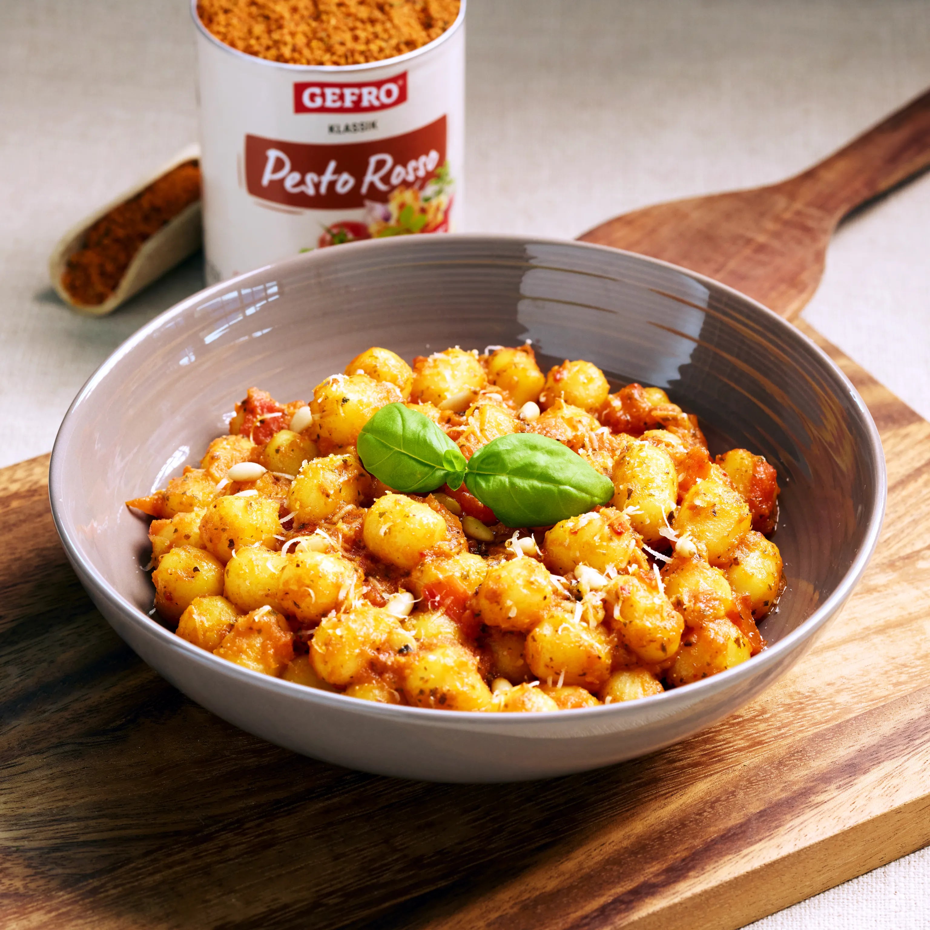 Schale mit Gnocchi in roter Pesto-Soße, garniert mit Basilikum und Käse, daneben Gewürzdose auf Holzbrett