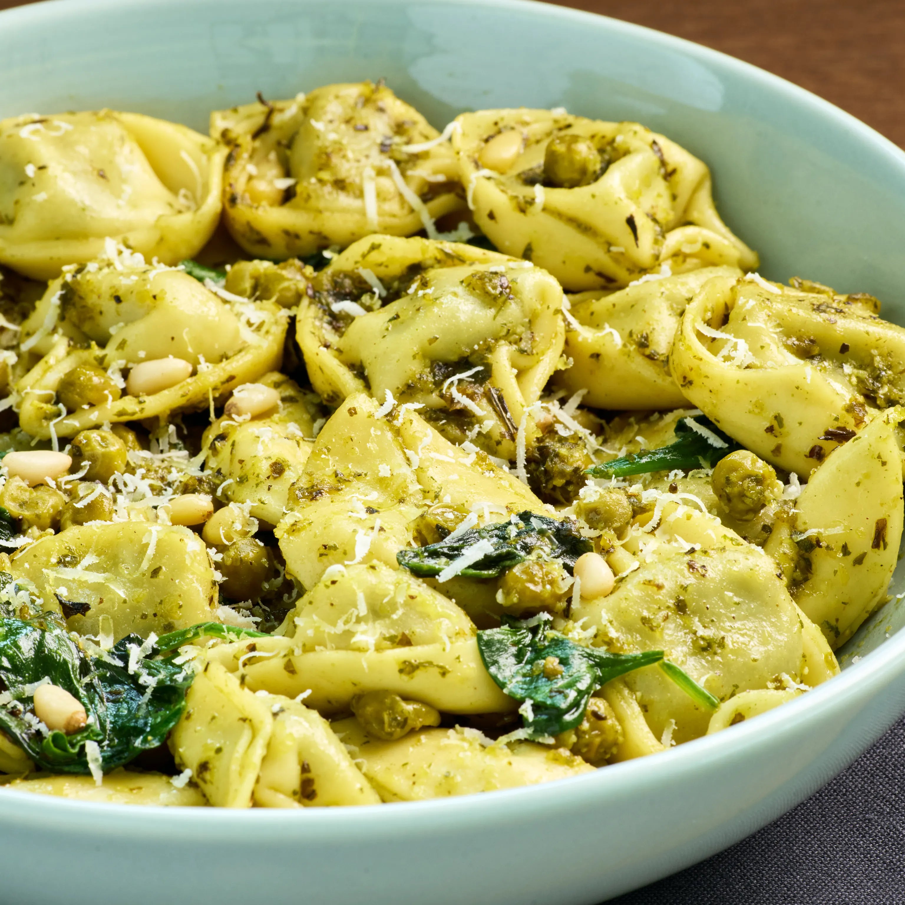 Tortellini mit grünem Pesto, Spinat, Kapern, Pinienkernen und geriebenem Käse in einer hellblauen Schale