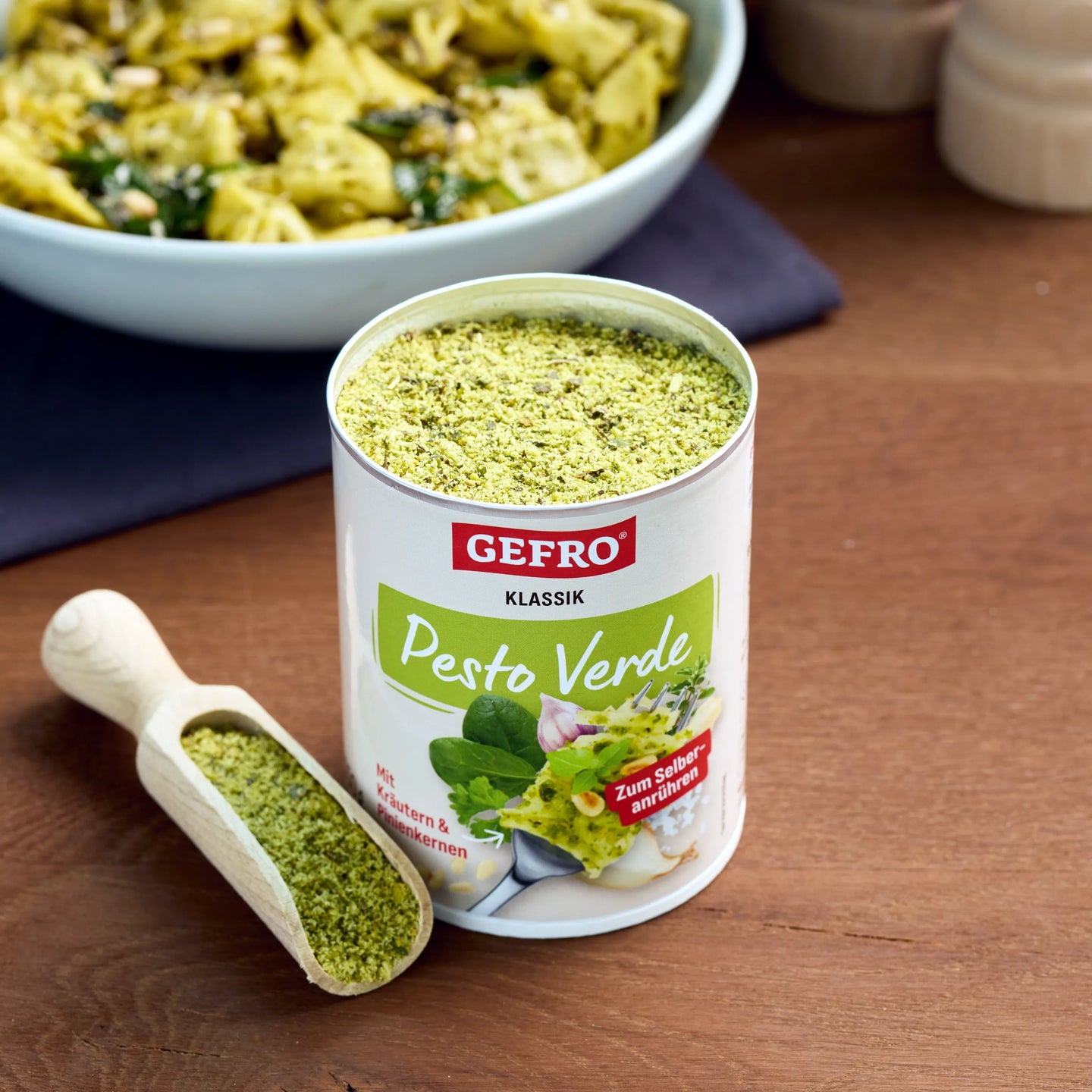 Dose GEFRO Pesto Verde mit grünem Pulver und Holzschaufel auf Holztisch, im Hintergrund eine Schüssel mit Pasta