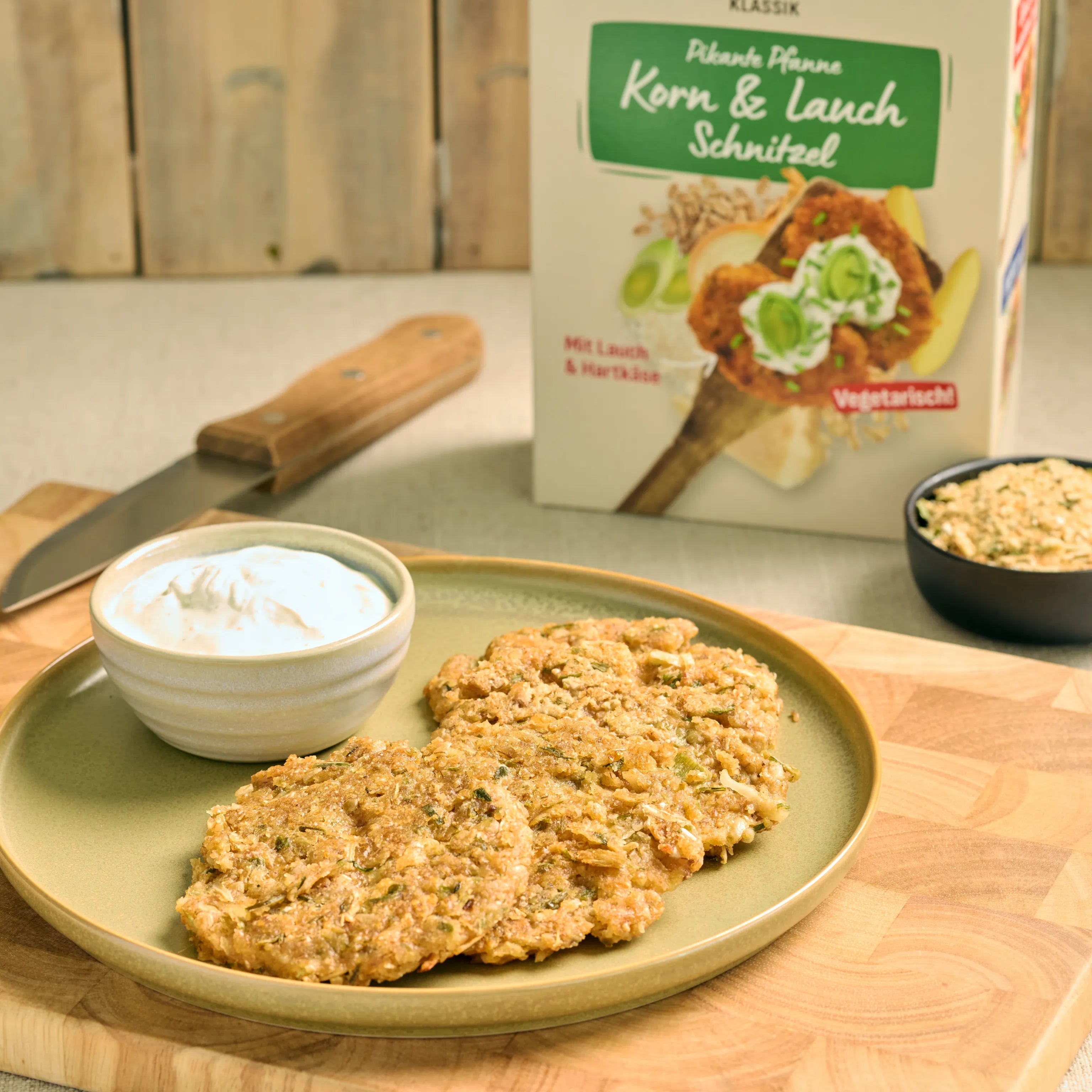 Zwei vegane Korn- und Lauch-Schnitzel auf einem Teller mit Dip, daneben die Verpackung auf einer Küchenarbeitsplatte