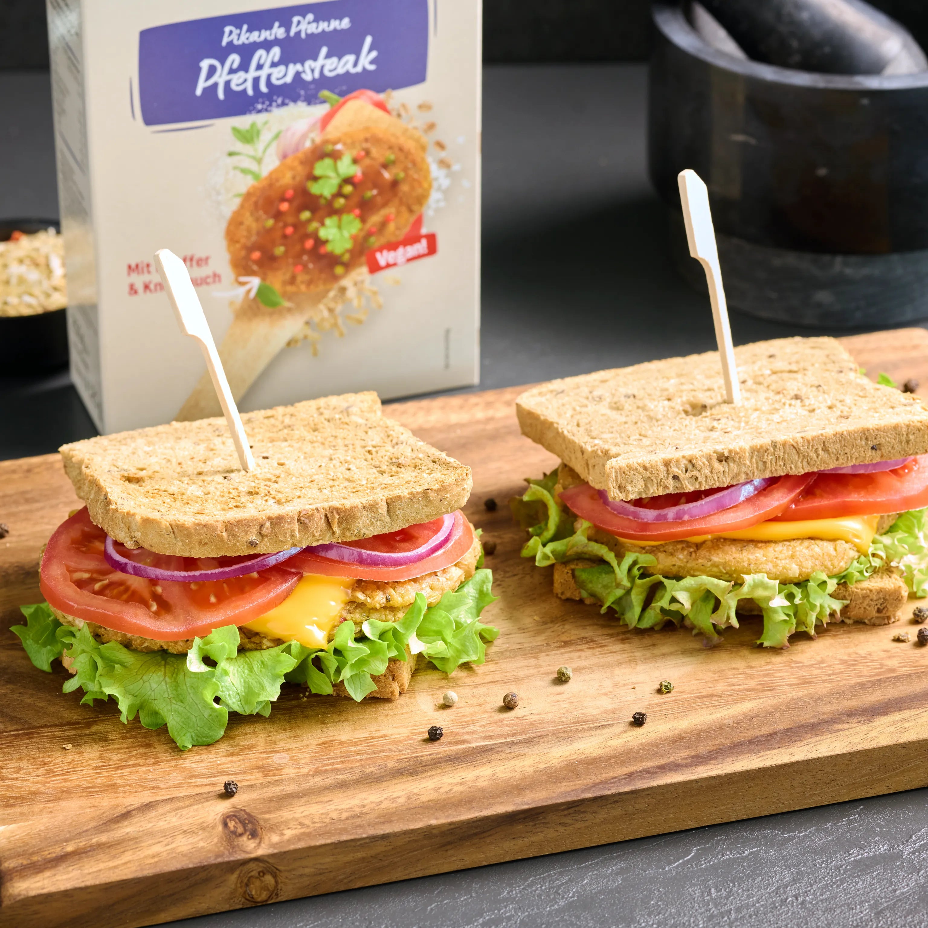 Zwei belegte Sandwiches mit Salat, Tomate, Zwiebeln und veganem Pfeffersteak auf einem Holzbrett serviert