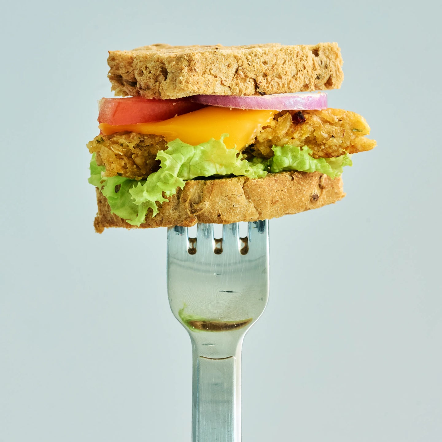 Sandwich mit Salat, Käse, Tomate und Zwiebel auf einer Gabel vor hellem Hintergrund, Nahaufnahme eines Snacks
