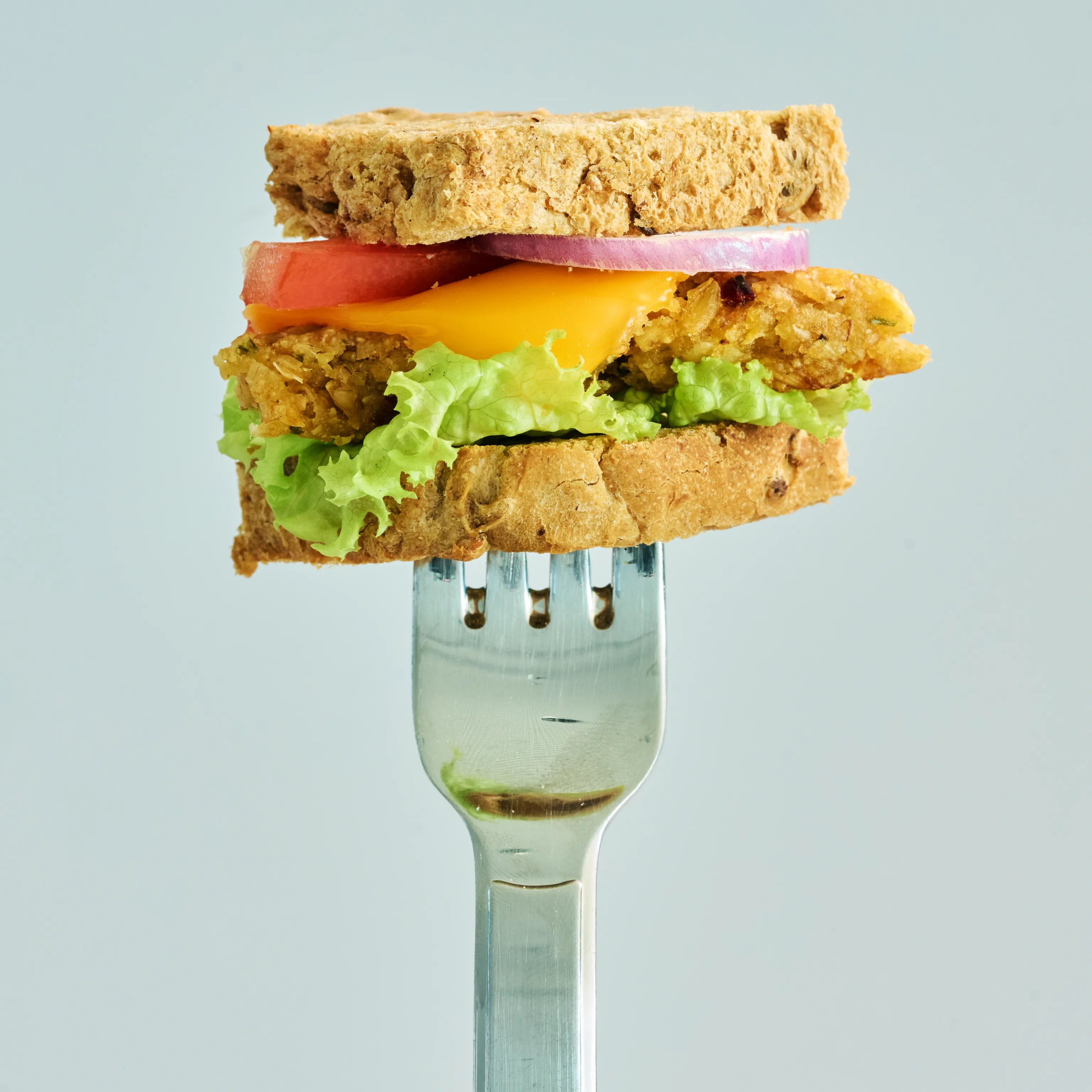 Sandwich mit Salat, Käse, Tomate und Zwiebel auf einer Gabel vor hellem Hintergrund, Nahaufnahme eines Snacks
