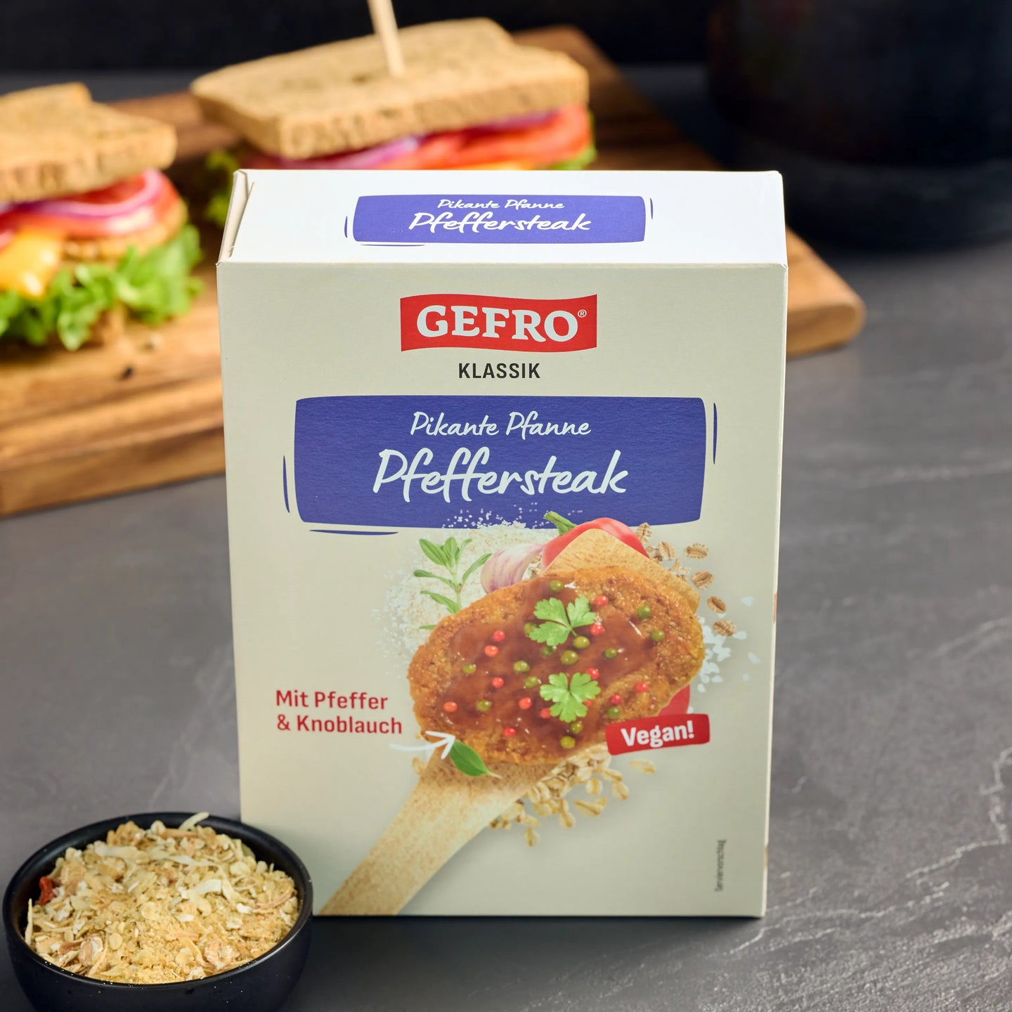 Verpackung von Gefro Klassik Pikante Pfanne Pfeffersteak, vegan, mit Pfeffer und Knoblauch, vor Sandwiches im Hintergrund