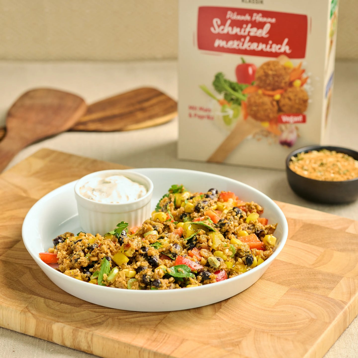 Schale mit mexikanischer Gemüse-Pfanne und Dip auf Holzbrett, dahinter eine Packung veganes Schnitzel Mix.