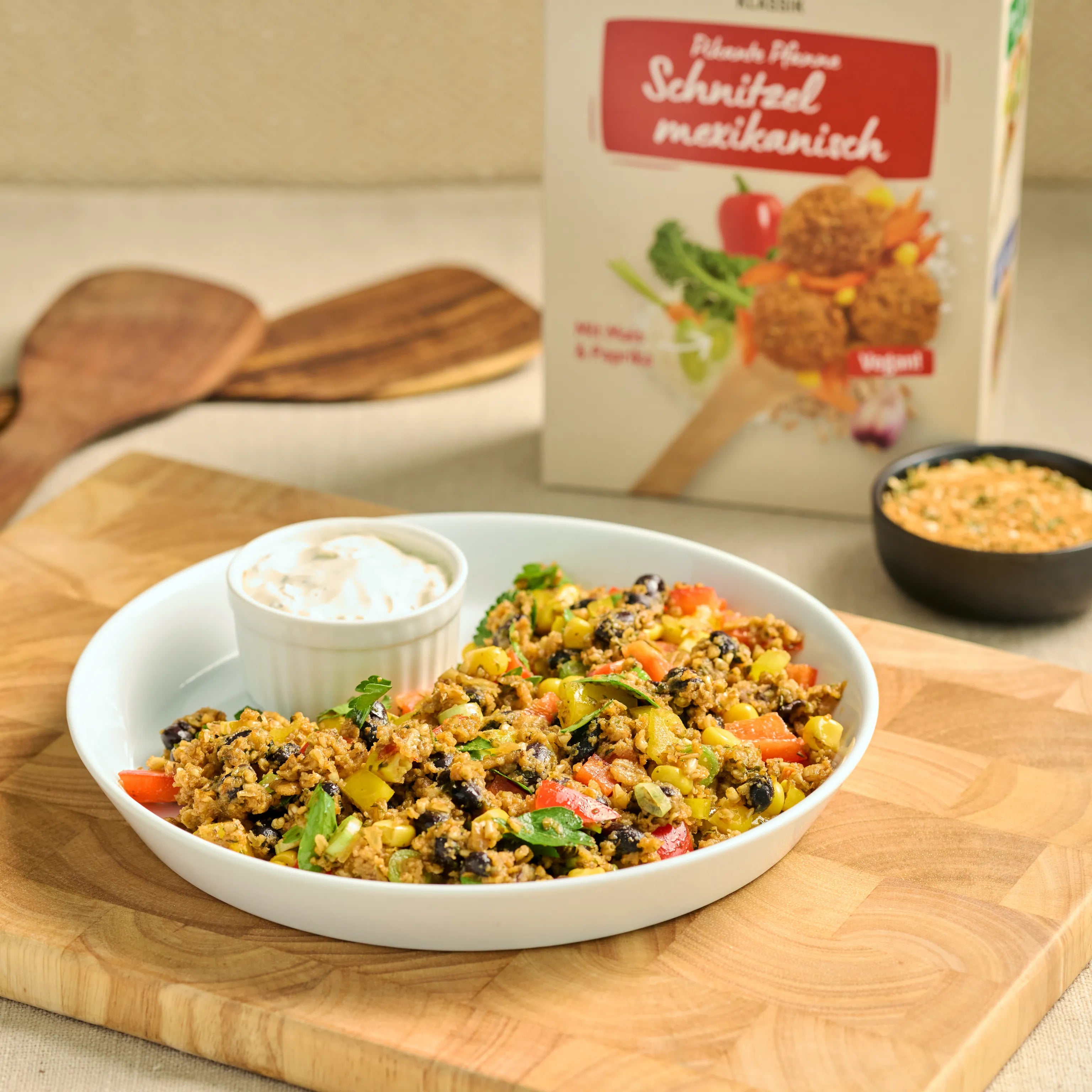 Schale mit mexikanischer Gemüse-Pfanne und Dip auf Holzbrett, dahinter eine Packung veganes Schnitzel Mix.