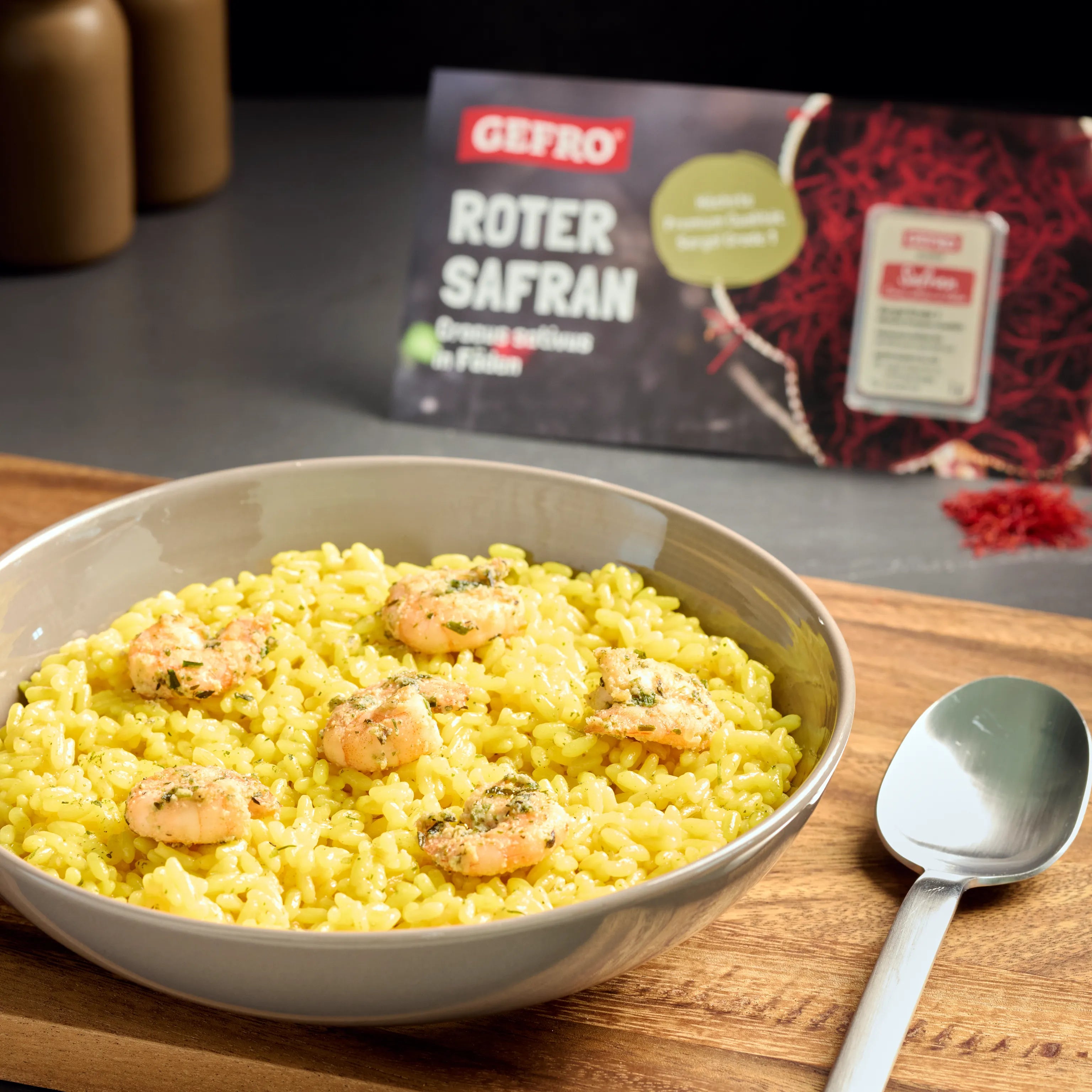 Schale mit gelbem Safranreis und Garnelen auf Holztisch, im Hintergrund Verpackung von Gefro Roter Safran.